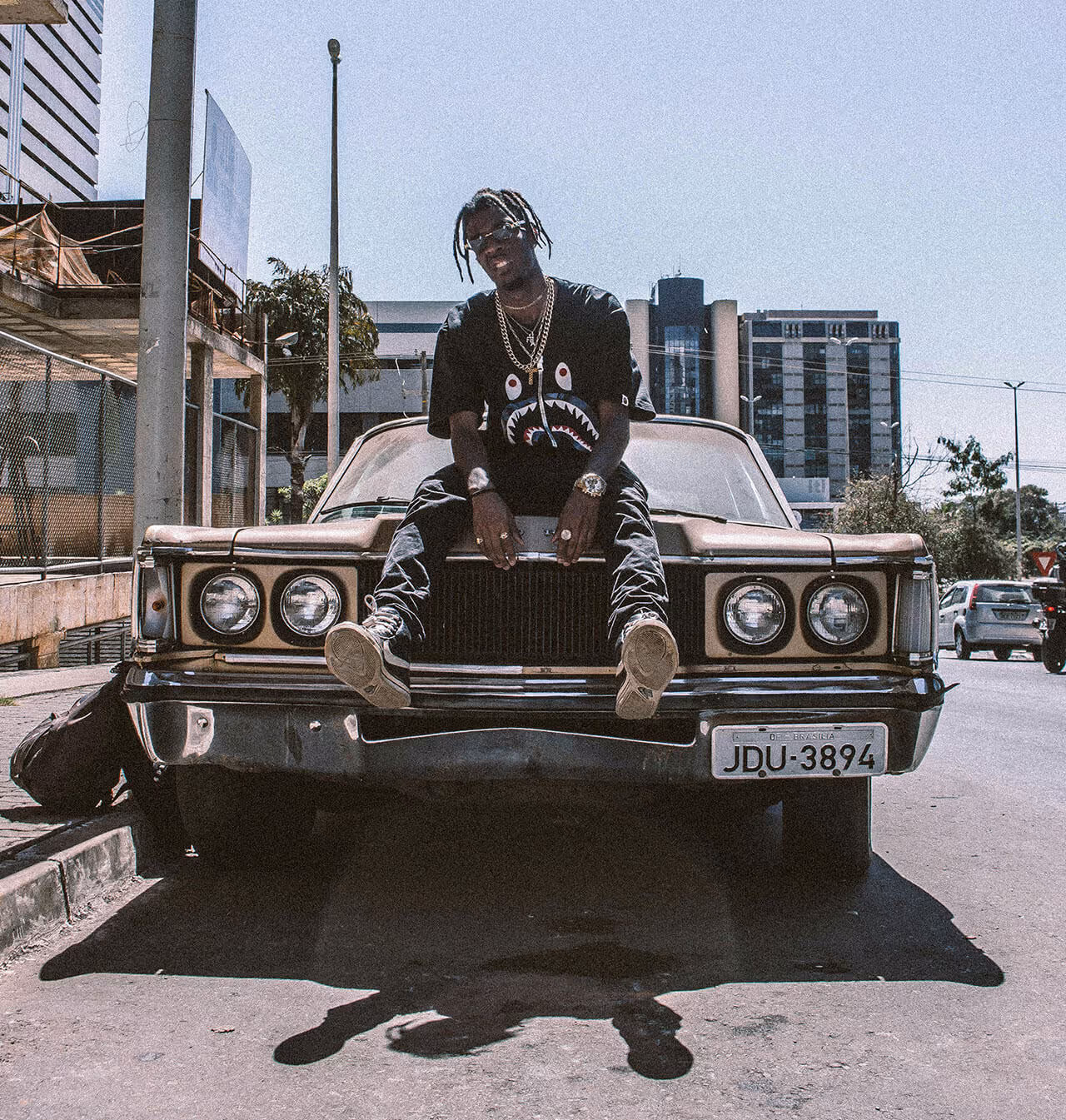 Urban streetwear fashion model sitting on vintage car in cityscape, showcasing hip-hop culture, gold chains, and retro style aesthetic.