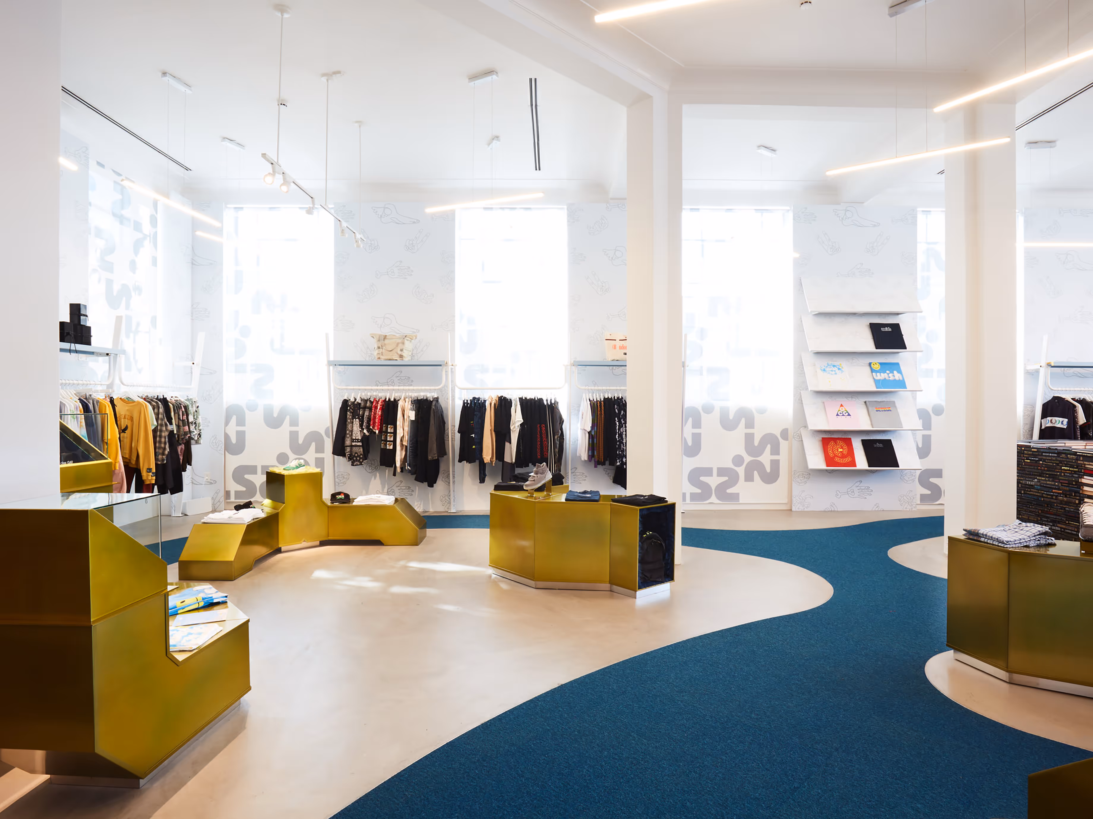 Interior of Wish ATL streetwear boutique showcasing a modern, minimalist retail design with curved blue carpeting, gold geometric display fixtures, and curated racks of designer clothing. T-shirts are neatly displayed on angled wall shelves, and natural light floods the space, highlighting urban fashion and high-end streetwear culture in Atlanta.