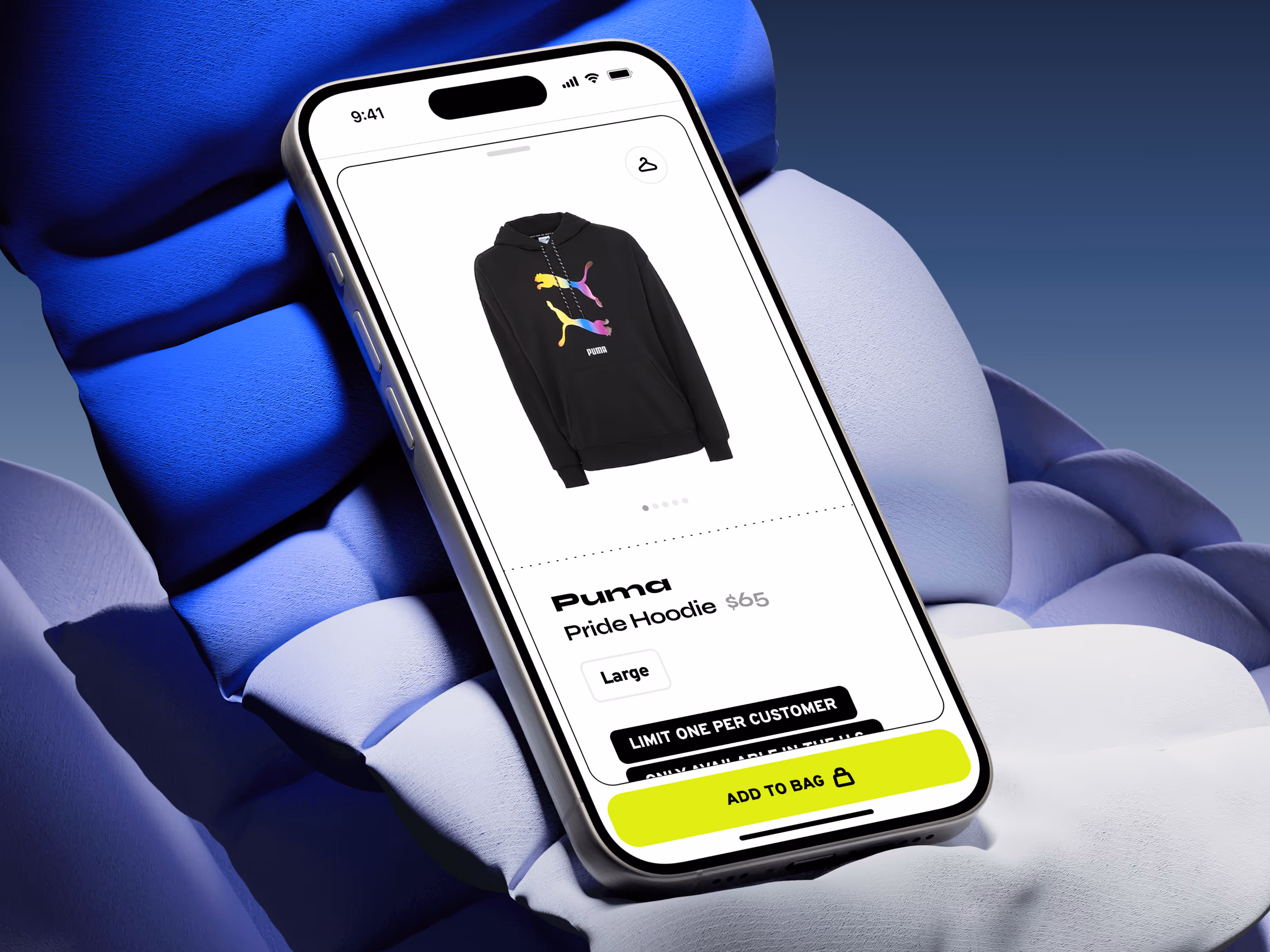 Smartphone displaying the Wish ATL mobile shopping interface for the Puma Pride Hoodie, featuring a multicolor pouncing cat graphic on a black sweatshirt. The screen highlights the price, size selection, and an “Add to Bag” button with a notice for limited availability. Set against a sculptural blue background, the image emphasizes mobile-first streetwear e-commerce and exclusive sneaker culture drops.