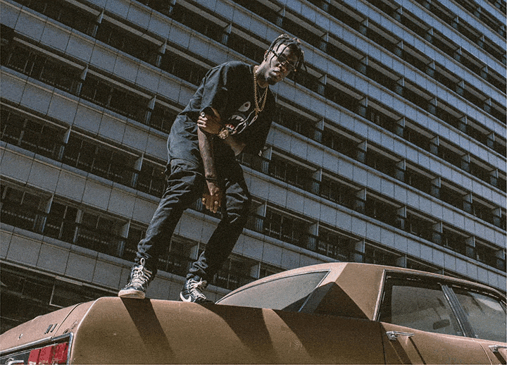 Streetwear fashion editorial shot of a man standing on top of a vintage brown car in an urban city setting. He wears a black graphic t-shirt, dark jeans, gold chains, and sneakers, posing confidently in front of a modern high-rise building. Captures hip-hop culture, urban lifestyle, and street style photography aesthetics.