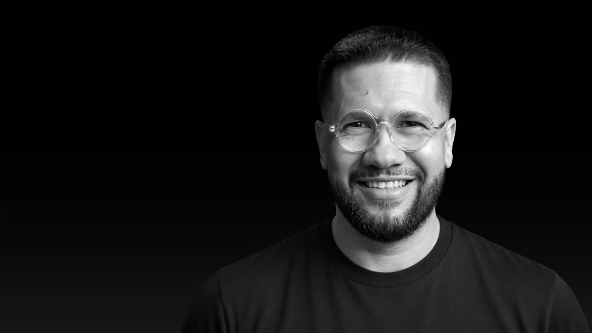Jesse smiling with short hair, beard, and round glasses against a black background.