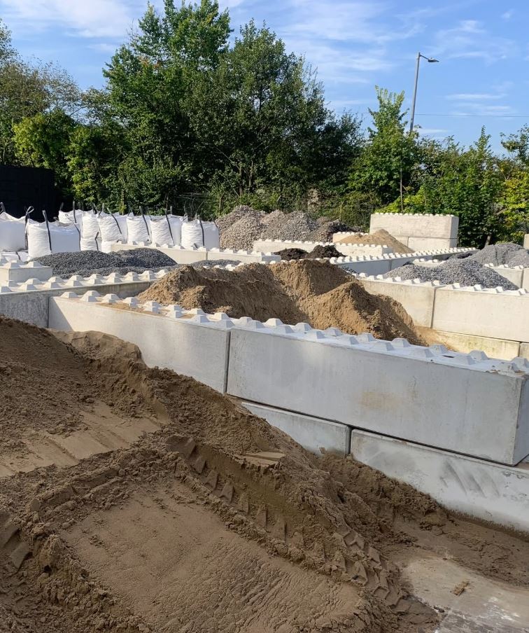 Construction site with multiple concrete block bays containing piles of sand, gravel, and other aggregate materials, with trees and blue sky in the background.
