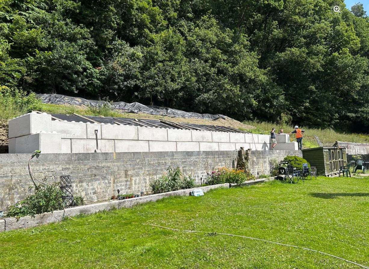 Construction site with a newly built concrete block retaining wall above an older stone wall, surrounded by green grass and dense trees in the background, with two workers standing on the wall and a garden shed and chairs nearby.