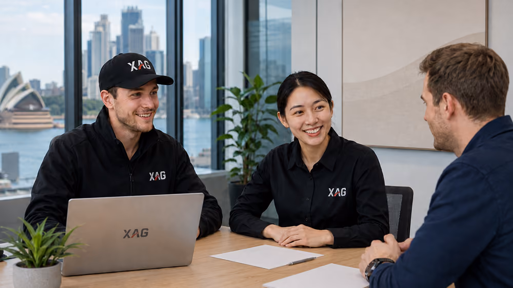 Three people smiling and talking at a table with laptops and papers in an office with city view.