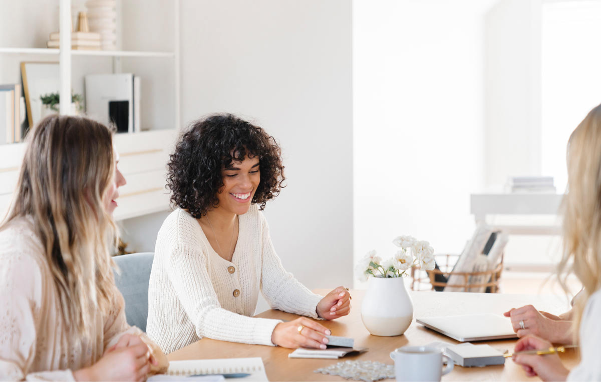 Three women sitting around a table engaged in a cheerful meeting with notebooks and a white vase with flowers.