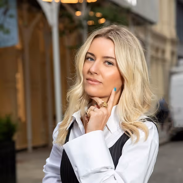 Portrait of a woman with blonde hair wearing a white shirt and black vest, photographed outdoors in a city environment.