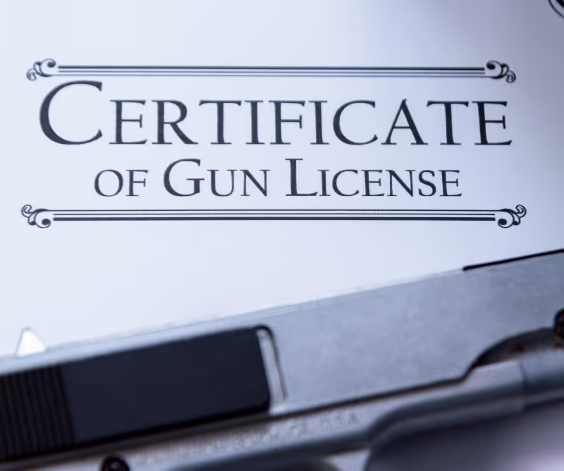 Close-up of a certificate titled 'Certificate of Gun License' with the edge of a handgun visible at the bottom.