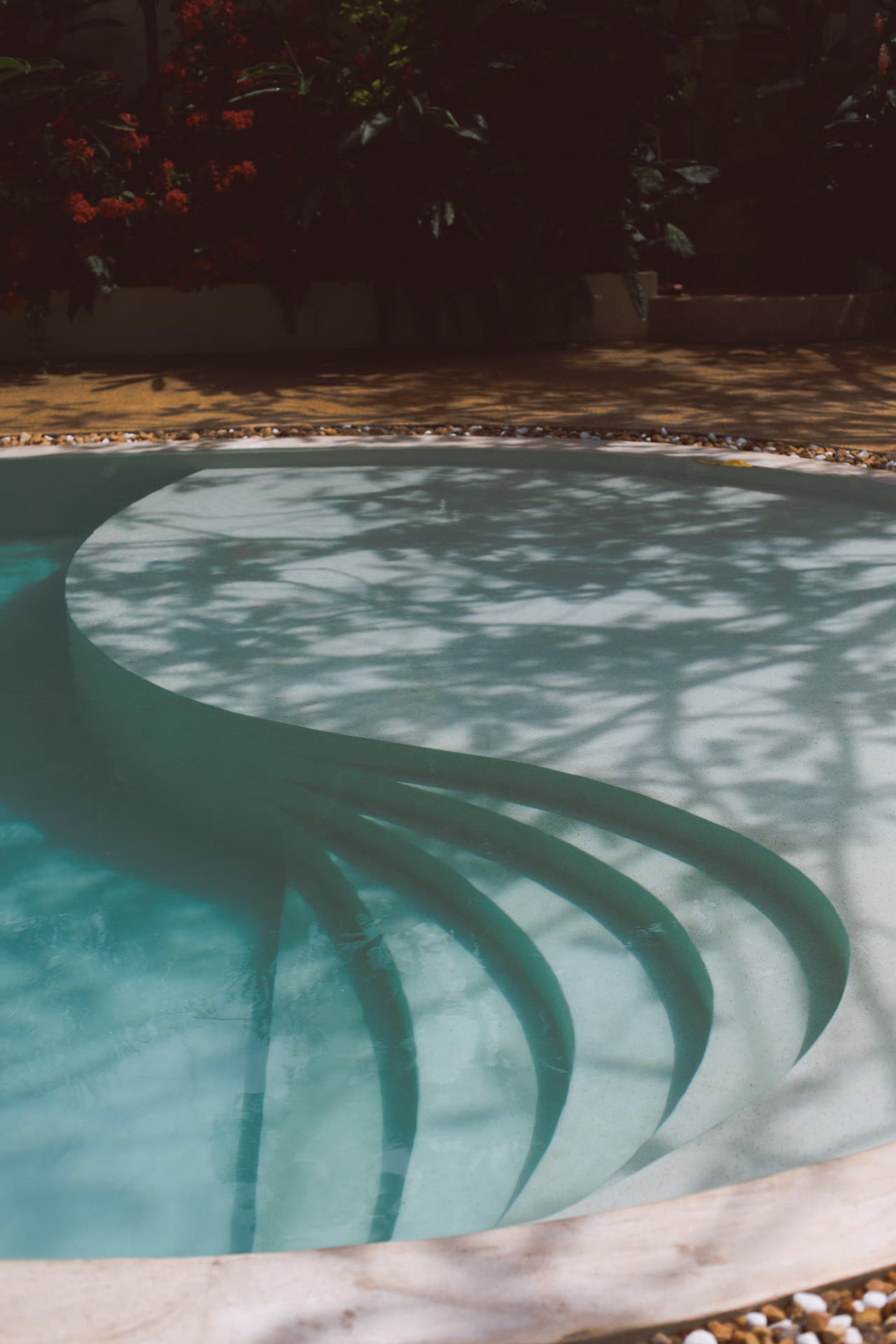 Hellblauer Pool mit runden, geschwungenen Stufen und Schatten von Pflanzen im Wasser.