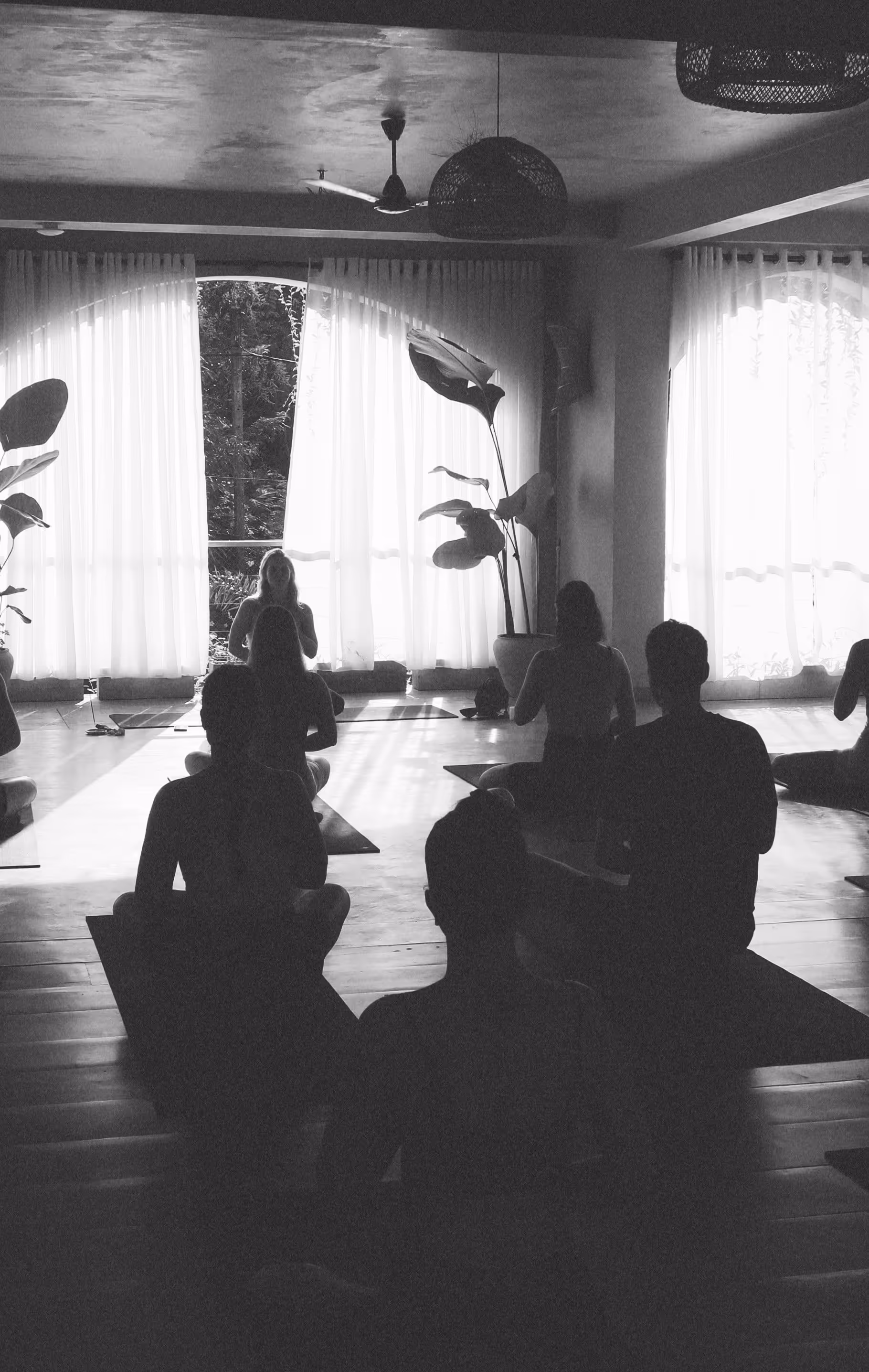Mehrere Personen sitzen im Meditationsraum mit großen Fenstern und weißen Vorhängen meditierend.