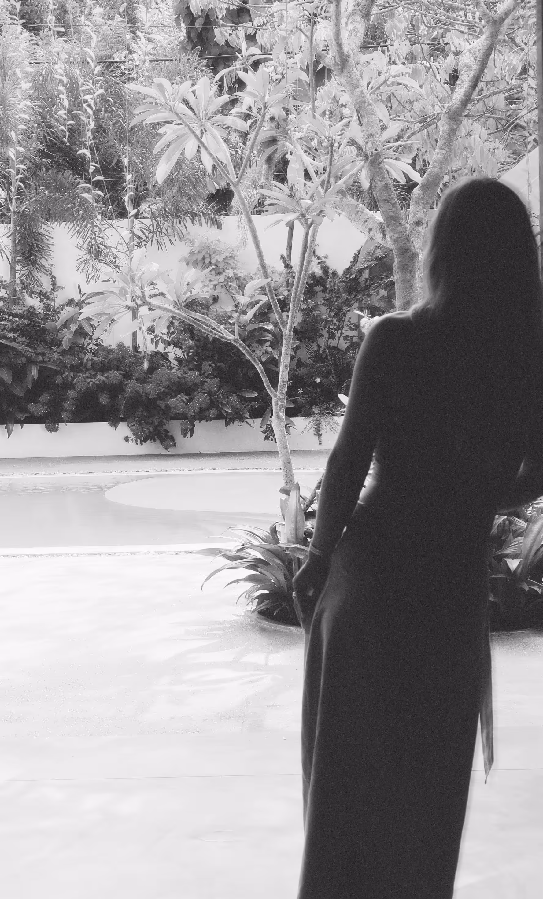 Silhouette einer Frau in langem Kleid, die auf einen Garten mit Pool und Bäumen blickt.
