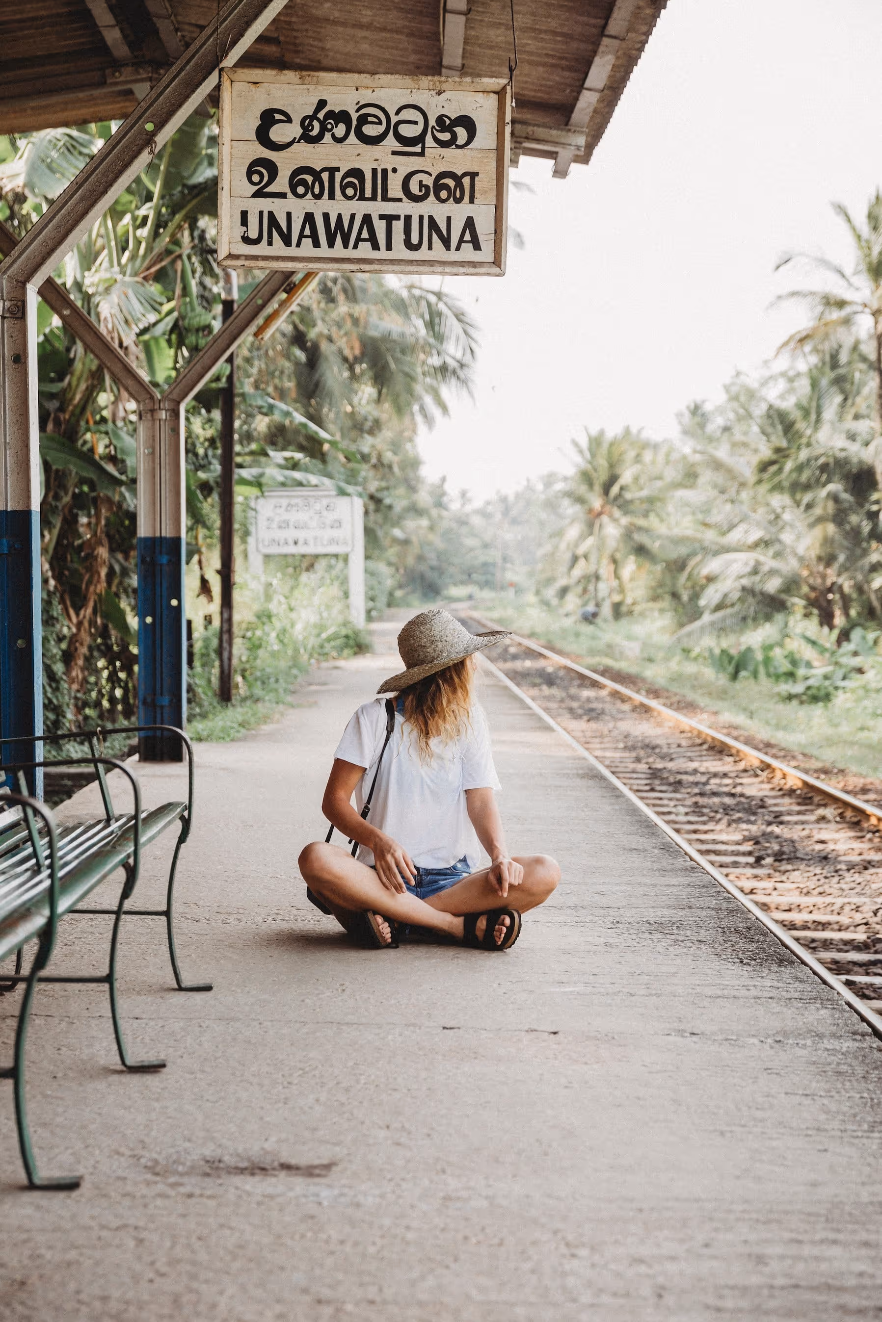 Frau mit Strohhut sitzt am Bahnsteig der Station Unawatuna neben Schienen und tropischer Vegetation.