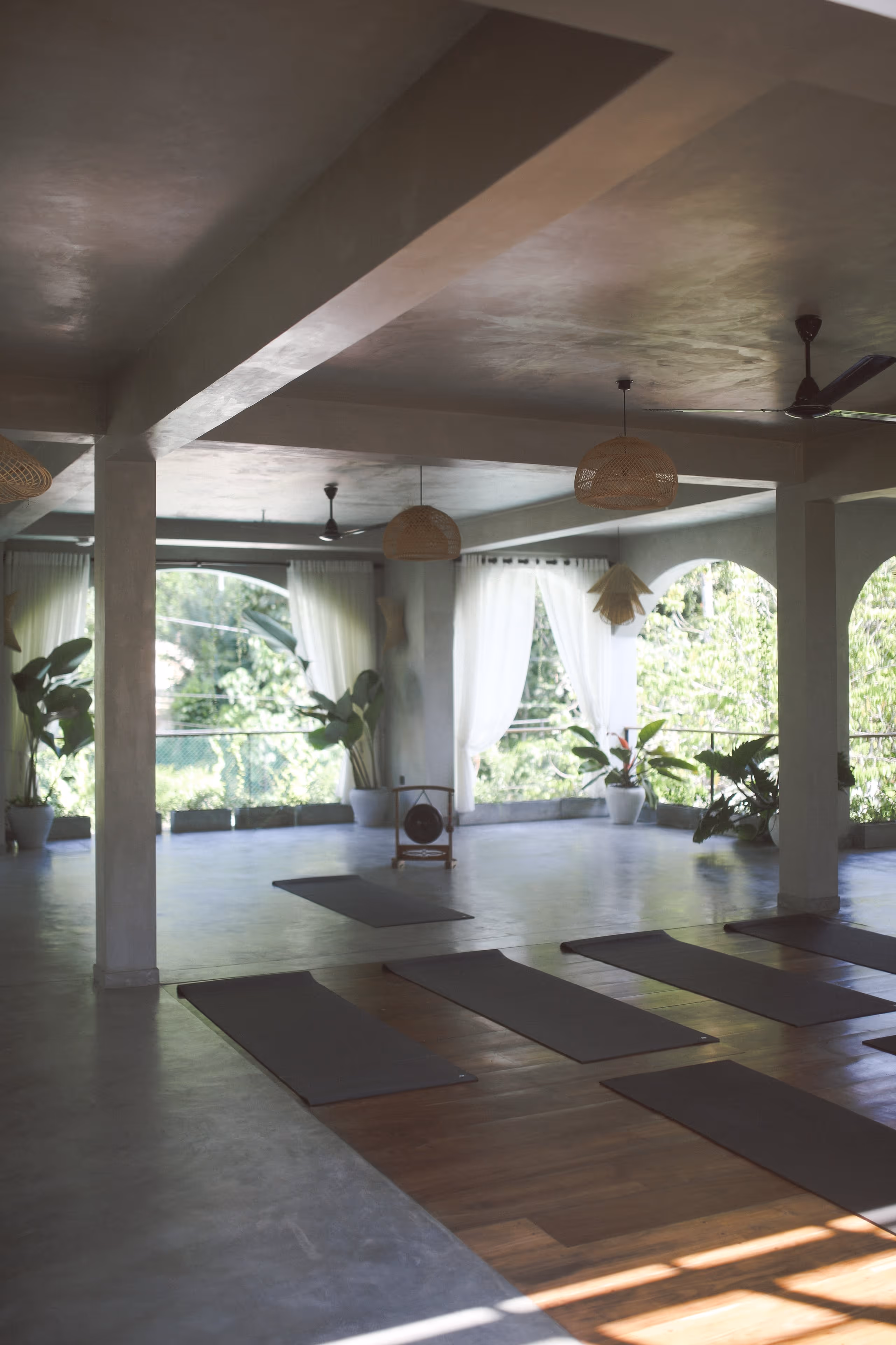 Heller Yoga-Raum mit Matten, großen Fenstern, Pflanzen und gewebten Lampenschirmen.