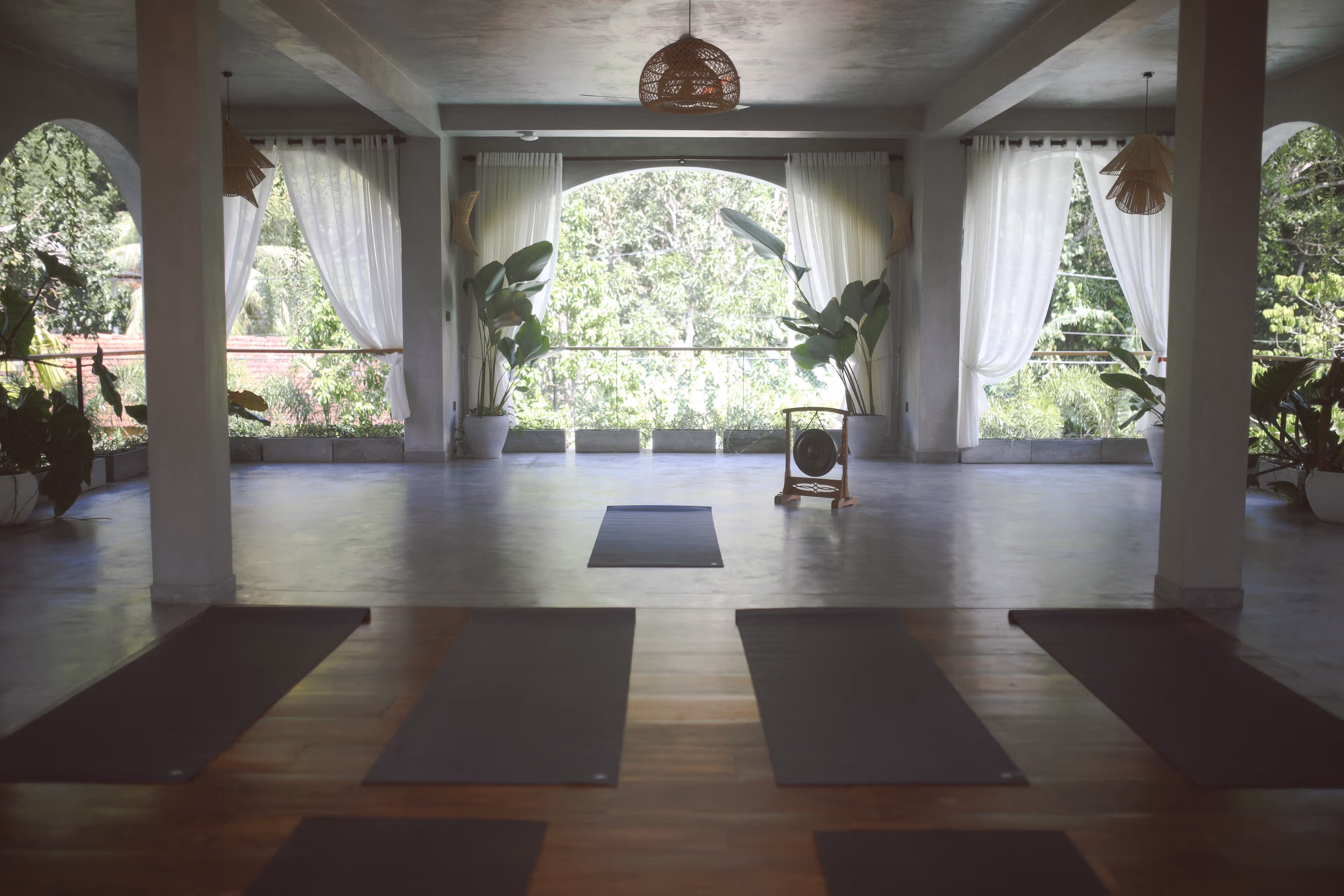 Yoga room with mats on wooden floor, large windows with sheer white curtains, plants, and a gong.
