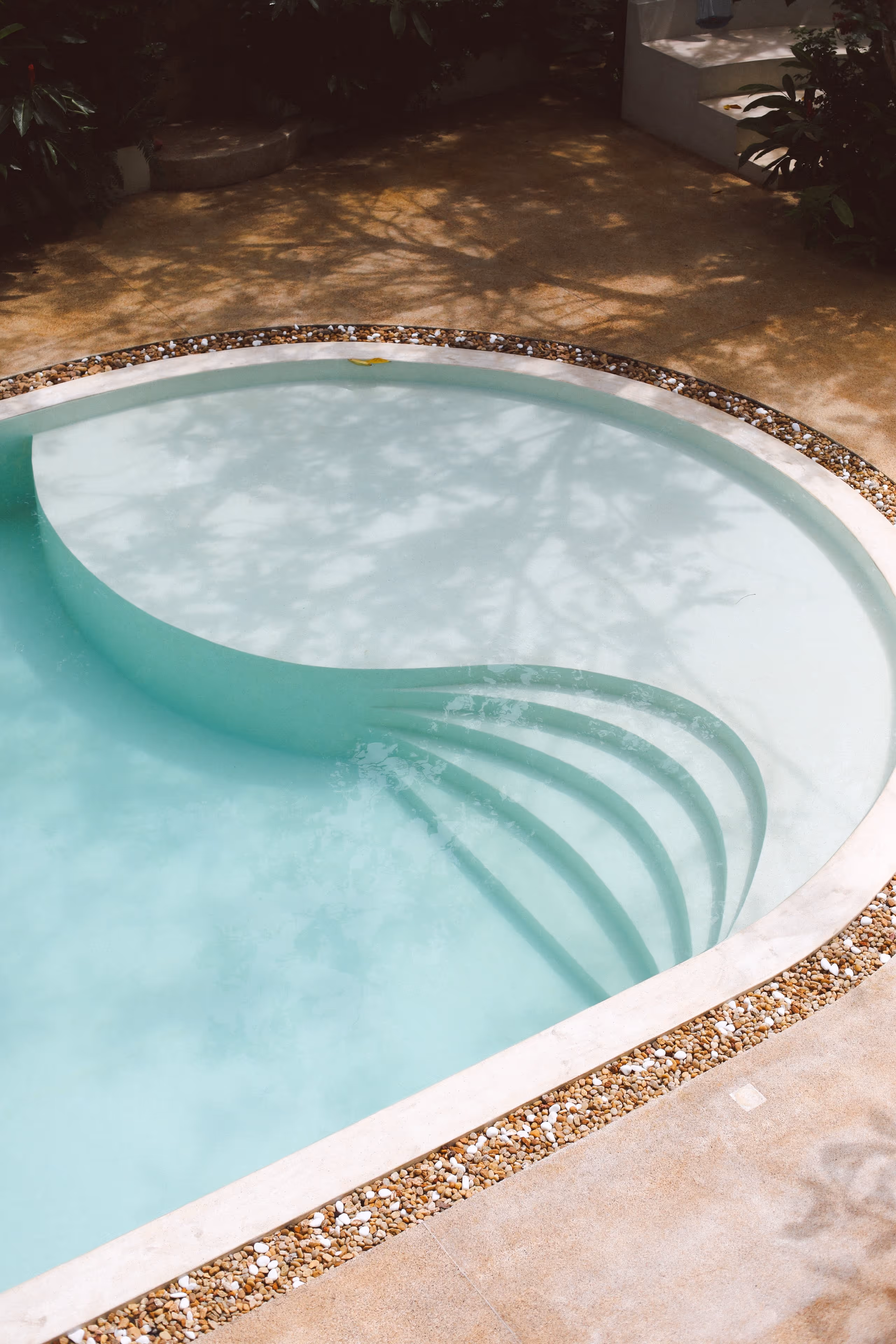 Rundes Schwimmbecken mit Stufen und Kies um den Rand, umgeben von Pflanzen und Schatten.