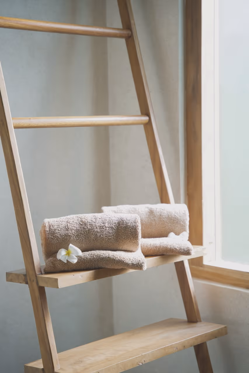 Zwei zusammengerollte Handtücher auf einem Holzregal neben einem Fenster und eine Blume.