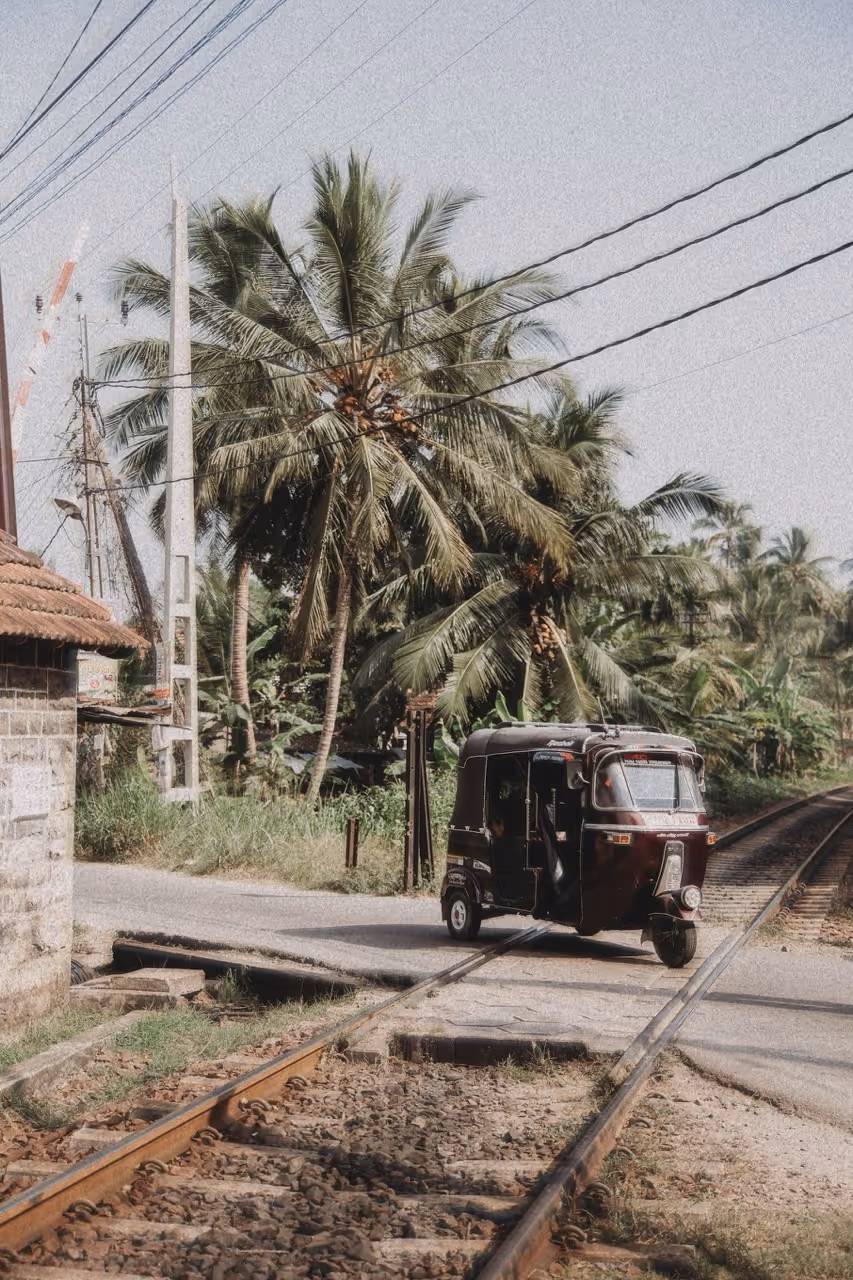 Ein schwarzer Tuk-Tuk-Fahrzeug fährt über eine Eisenbahnkreuzung mit Palmen im Hintergrund.