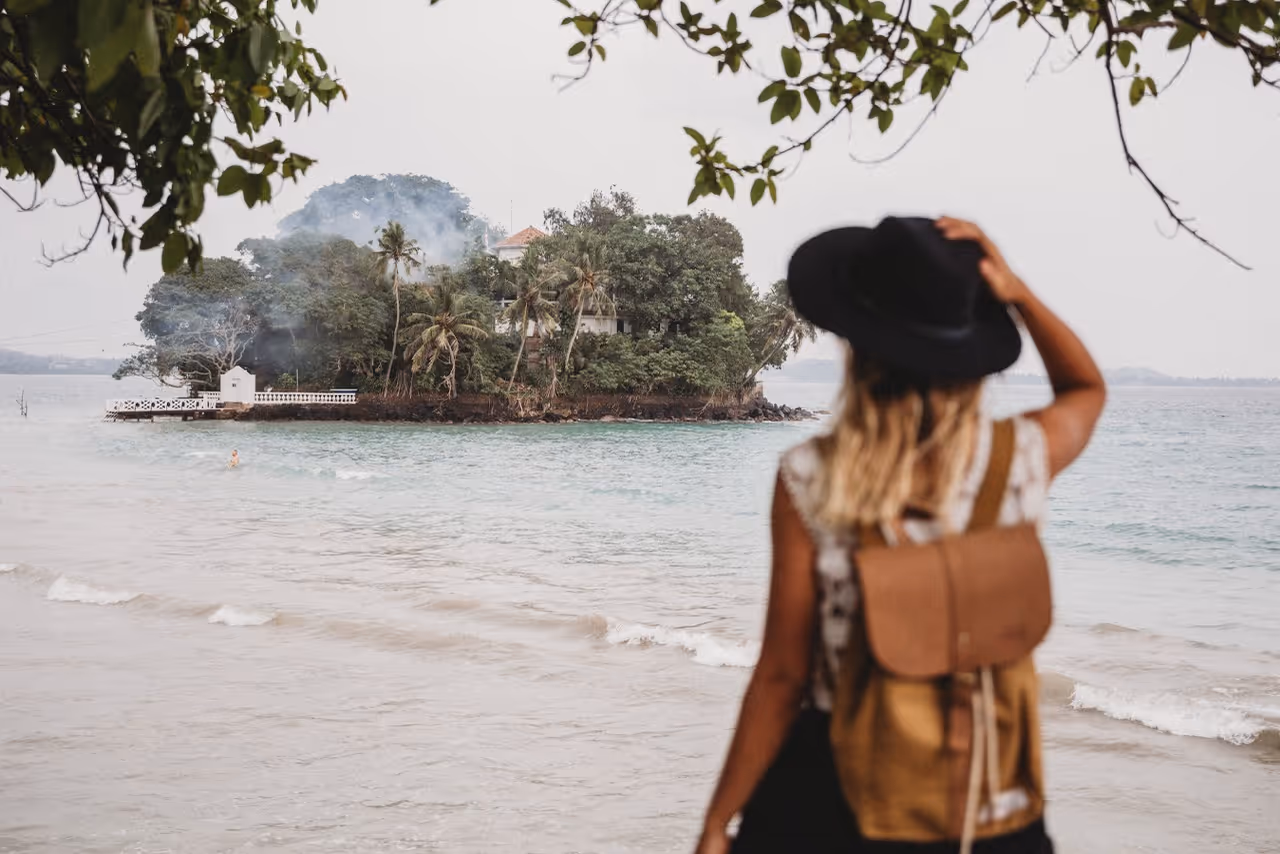 Frau mit Hut und Rucksack blickt auf bewaldete Insel im Meer vom Strand aus.
