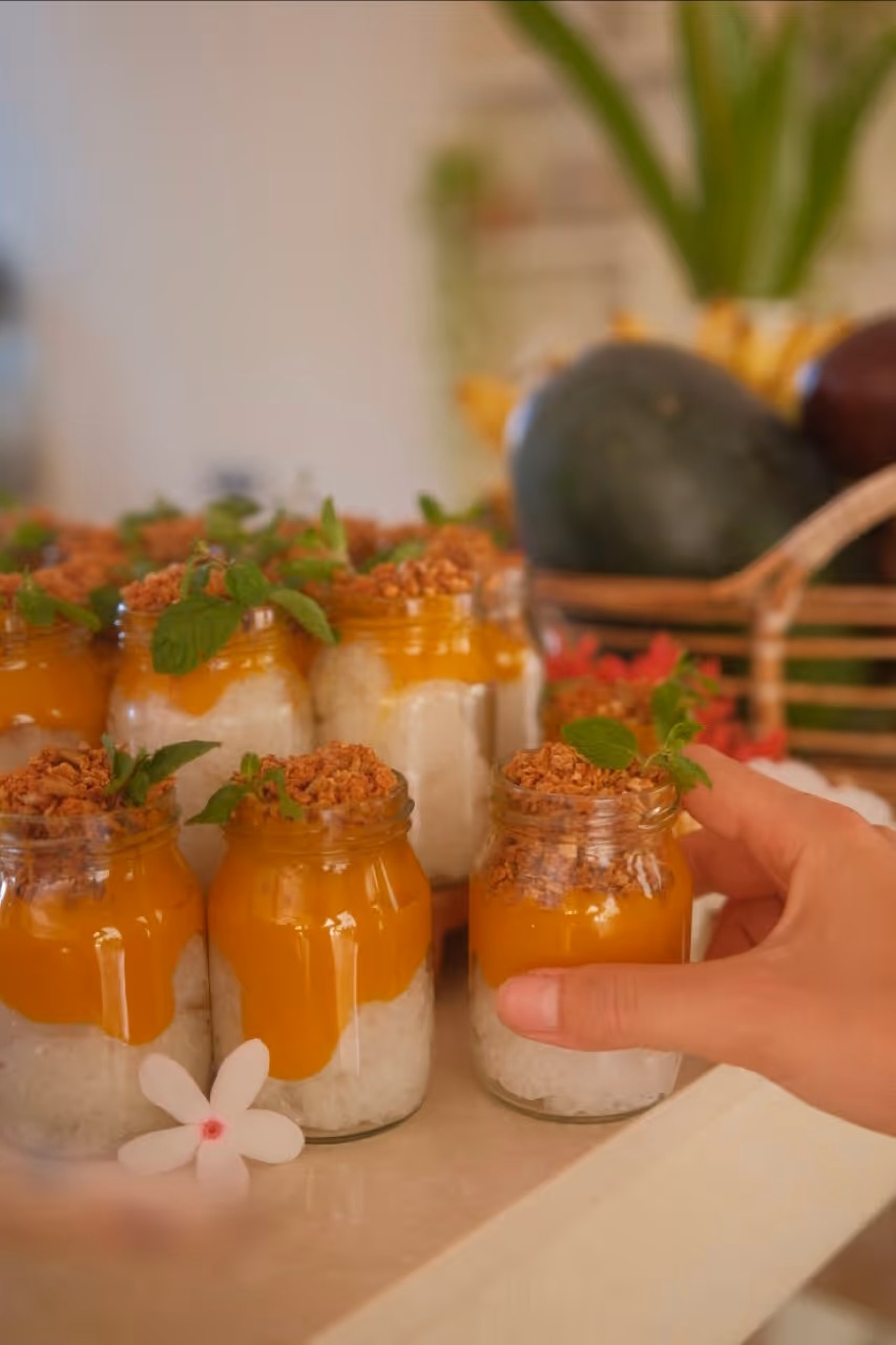 Hand nimmt ein Glas mit Schichtdessert aus Reis, Mangopüree und Granola, garniert mit Minze.