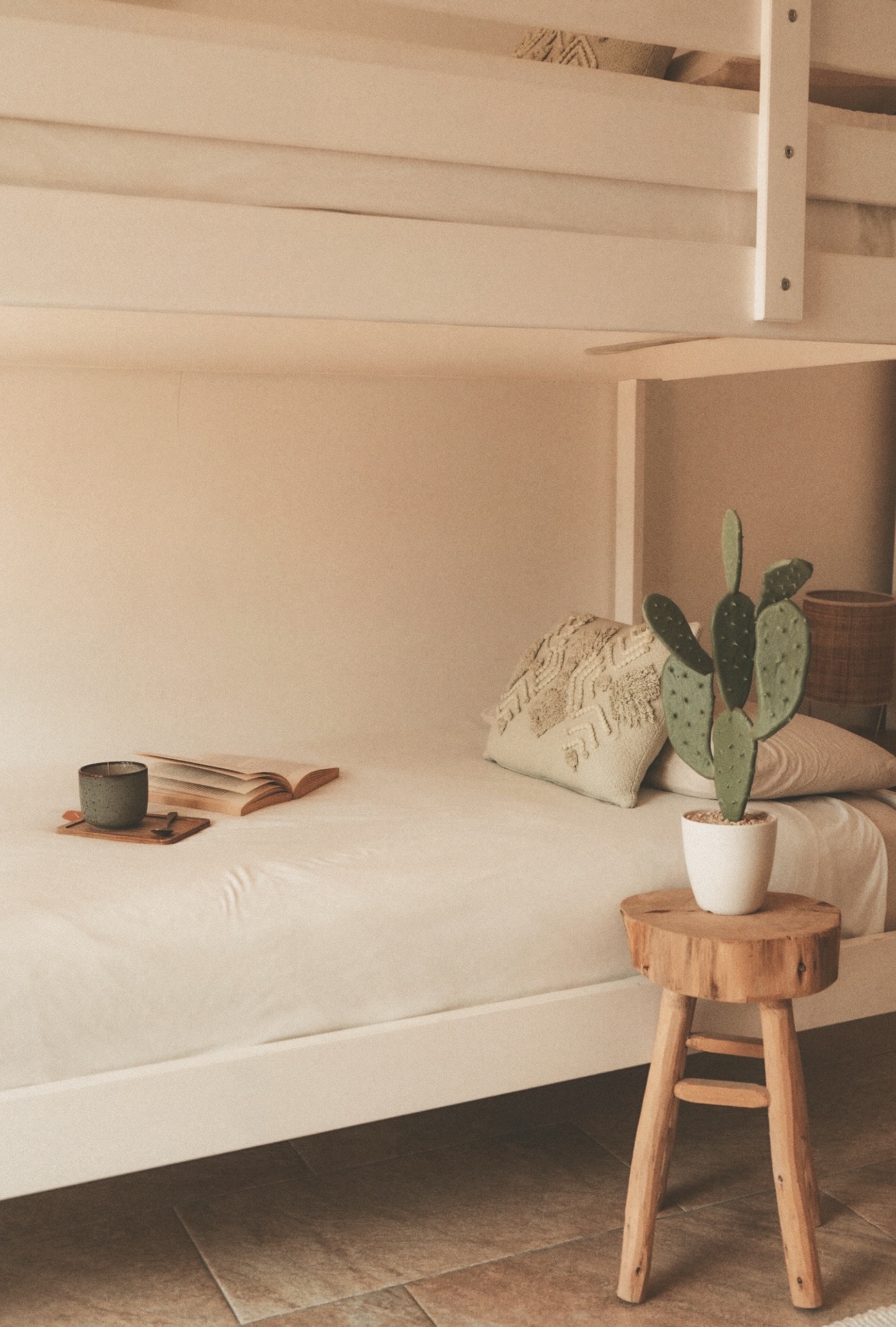 Gemütliches weißes Bett mit Kissen, offenem Buch und Kerze, daneben Holzstuhl mit Kaktus.