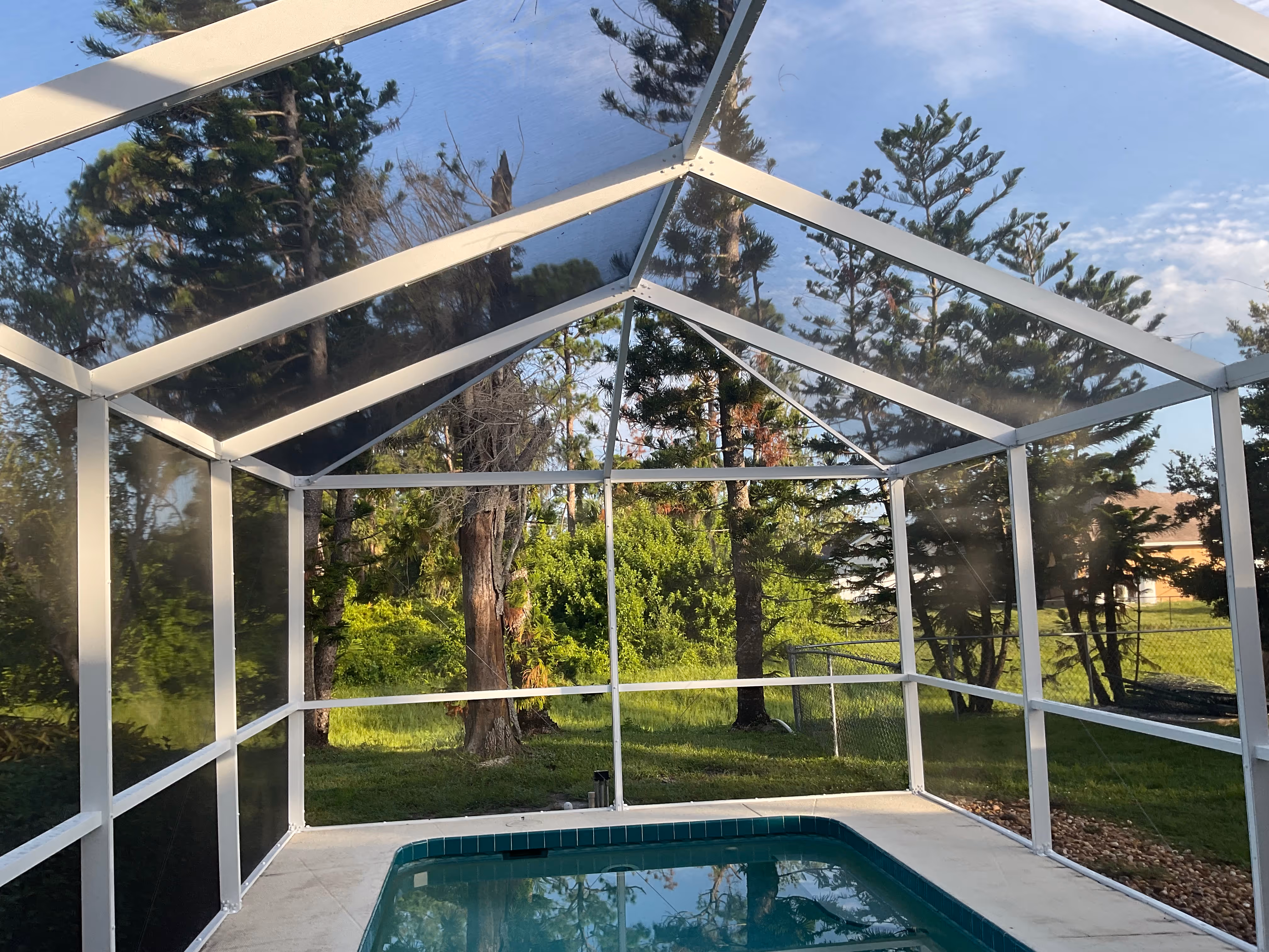 Interior view of white aluminum pool screen enclosure with translucent screen roof and pool, trees visible beyond — AB Aluminum & Screens