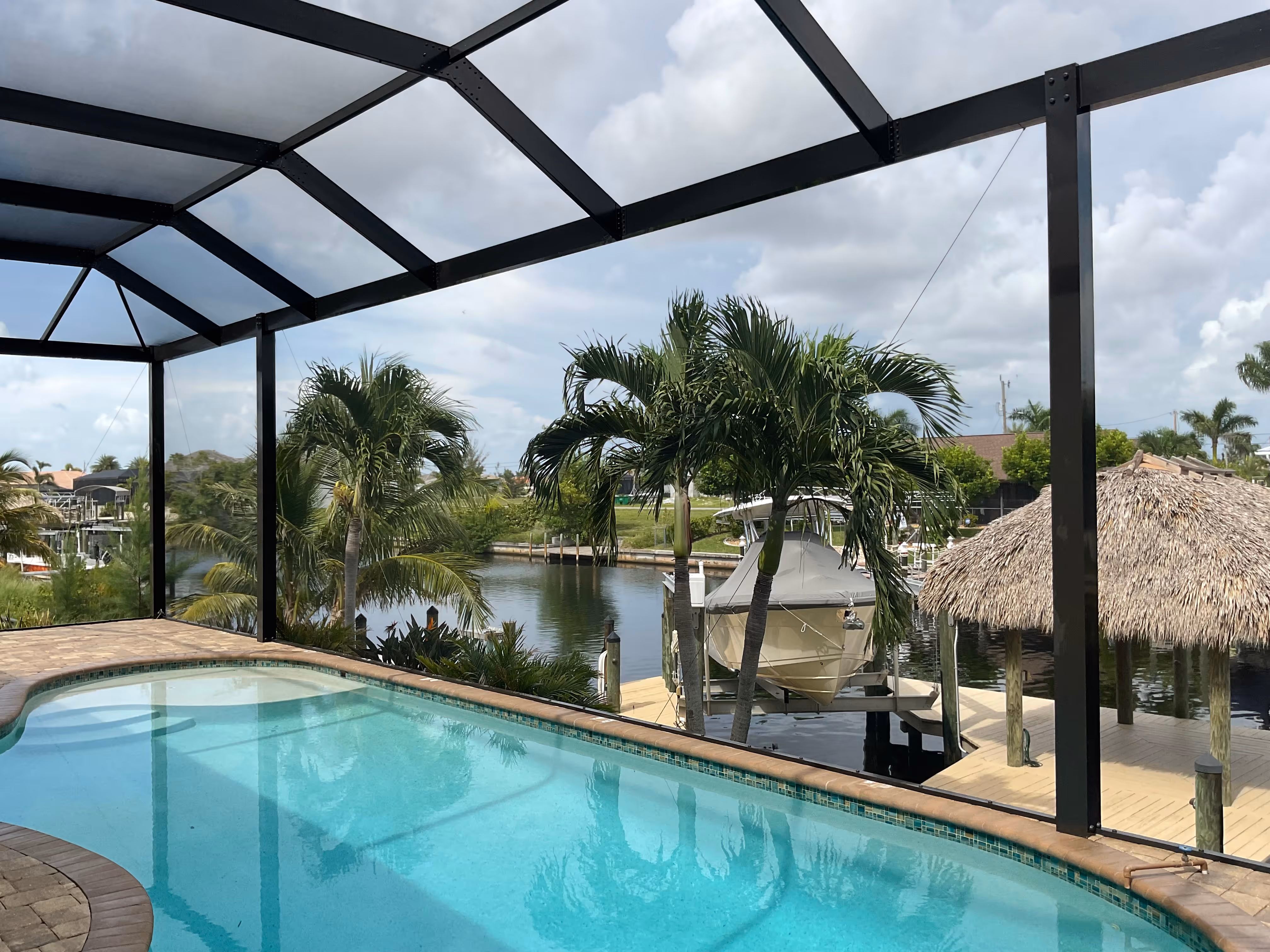 Interior view of dark bronze pool enclosure with irregular-shaped pool, waterfront canal with private boat dock, tiki hut and palm trees — AB Aluminum & Screens