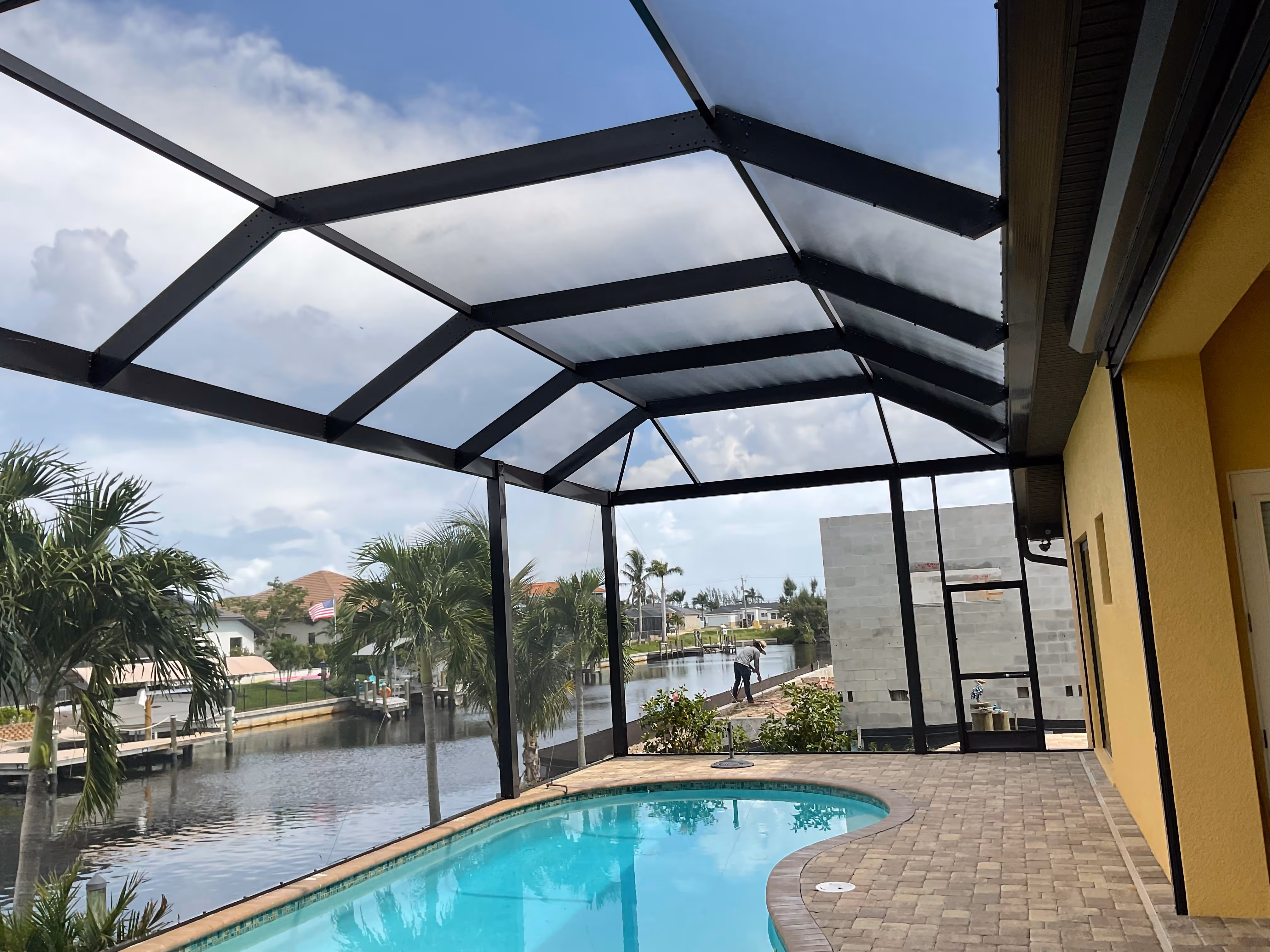 Dark bronze aluminum pool enclosure interior with curved pool and paver deck overlooking wide waterfront canal with docks, palm trees and luxury homes — AB Aluminum & Screens
