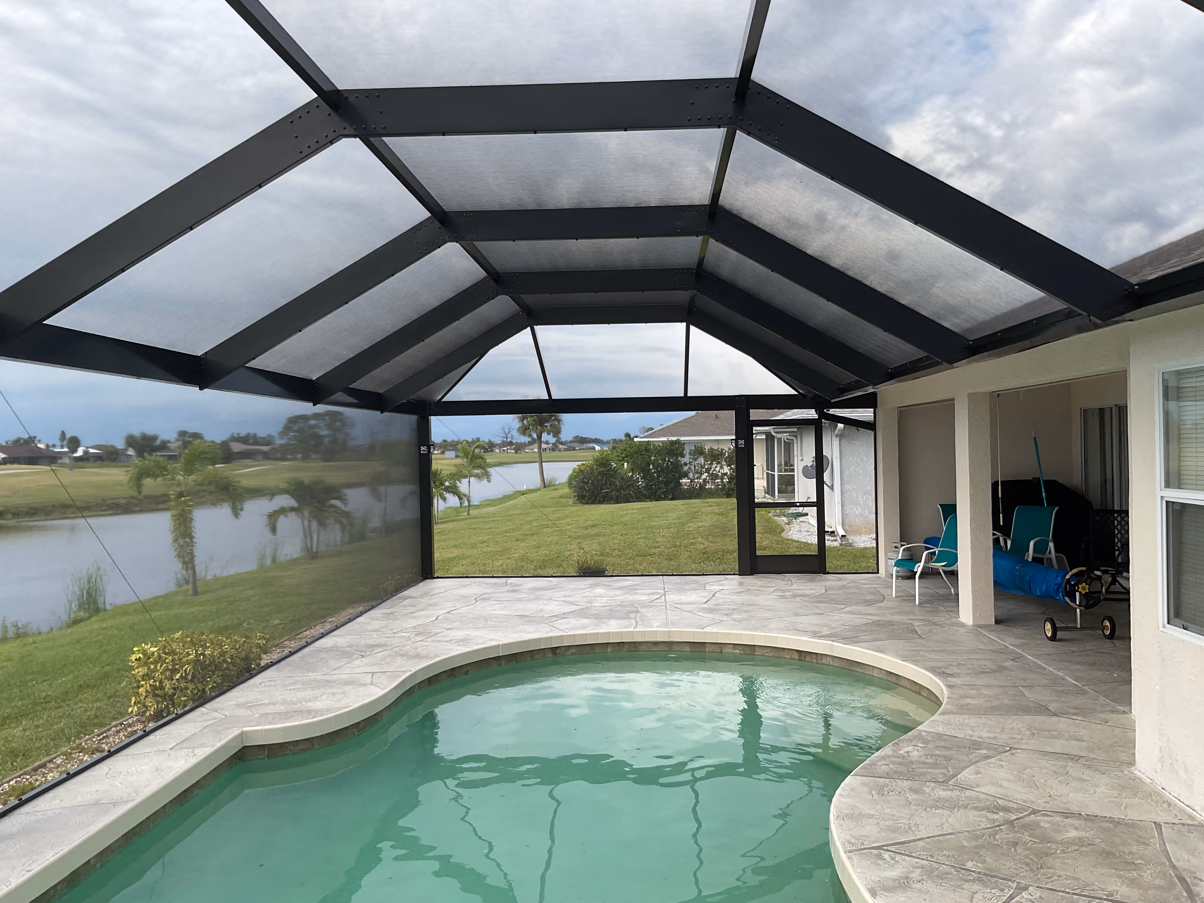 Dark bronze aluminum pool screen enclosure interior, freeform pool with open lake view, stamped concrete paver deck and patio furniture, South Florida — AB Aluminum & Screens