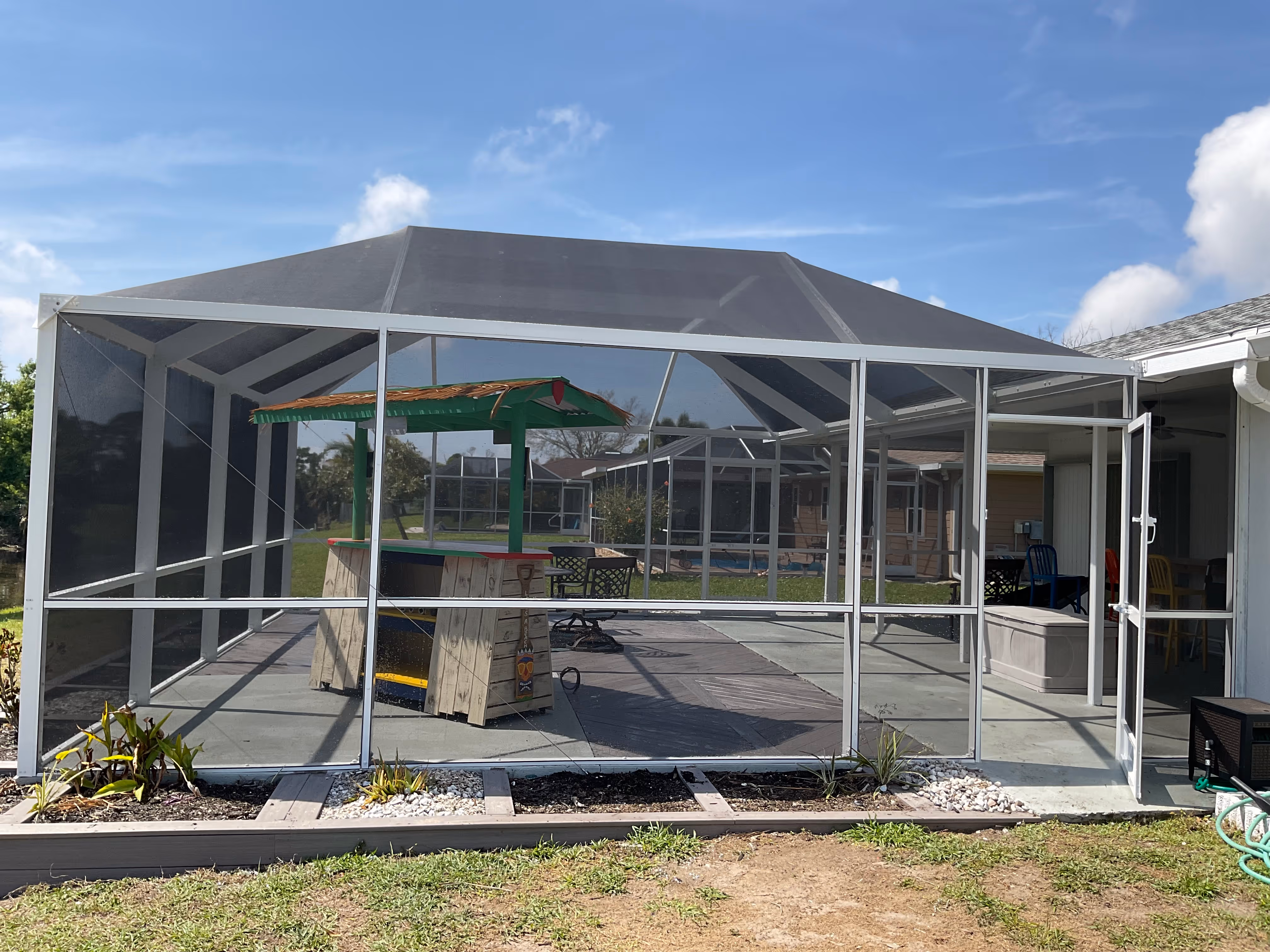 White aluminum pool screen enclosure exterior, completed gable roof, garden beds with tropical plants along base, green lawn, South Florida — AB Aluminum & Screens