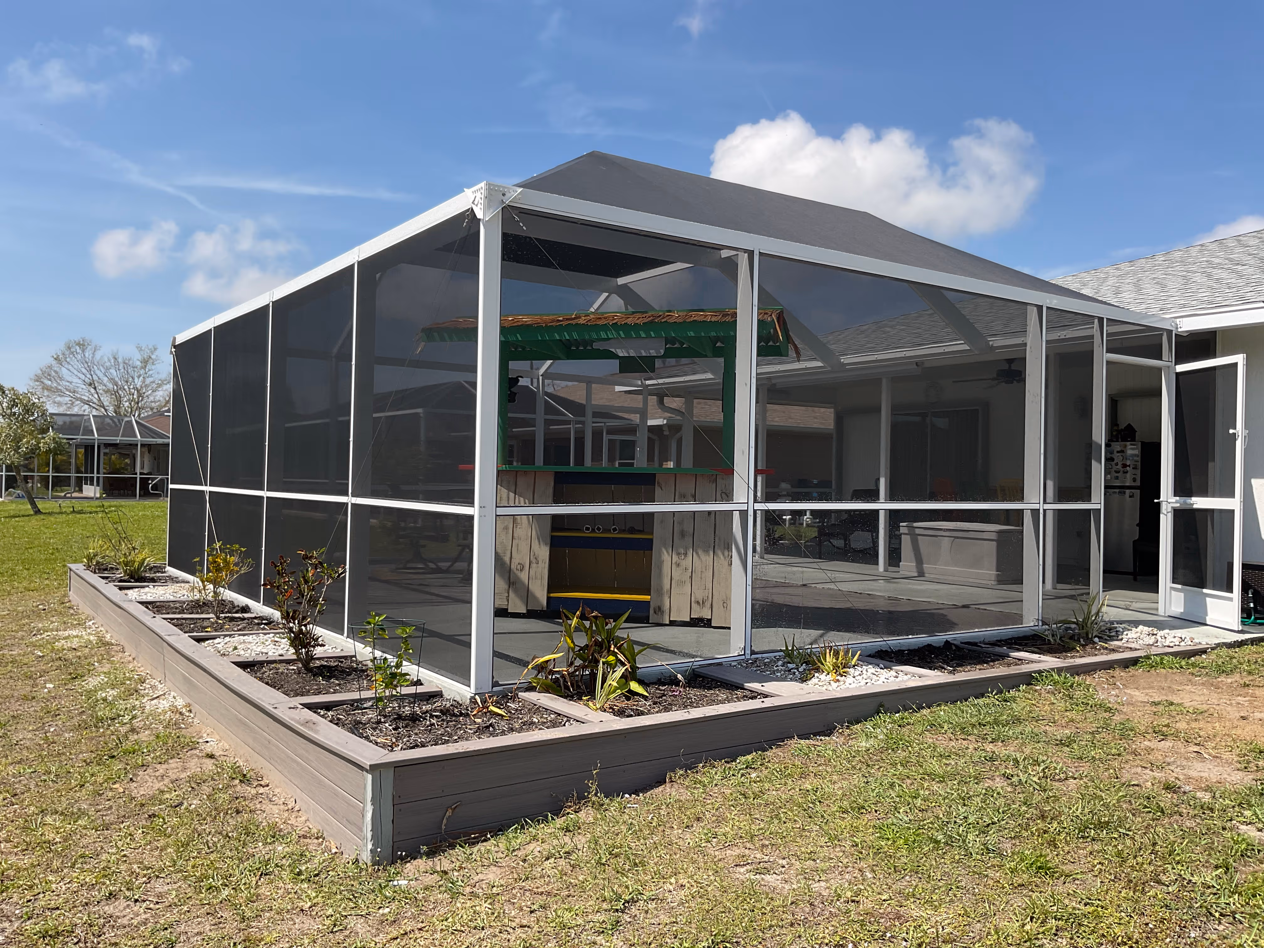 White aluminum pool screen enclosure exterior, freshly installed gable roof, tropical garden beds and green lawn, single-story South Florida home — AB Aluminum & Screens