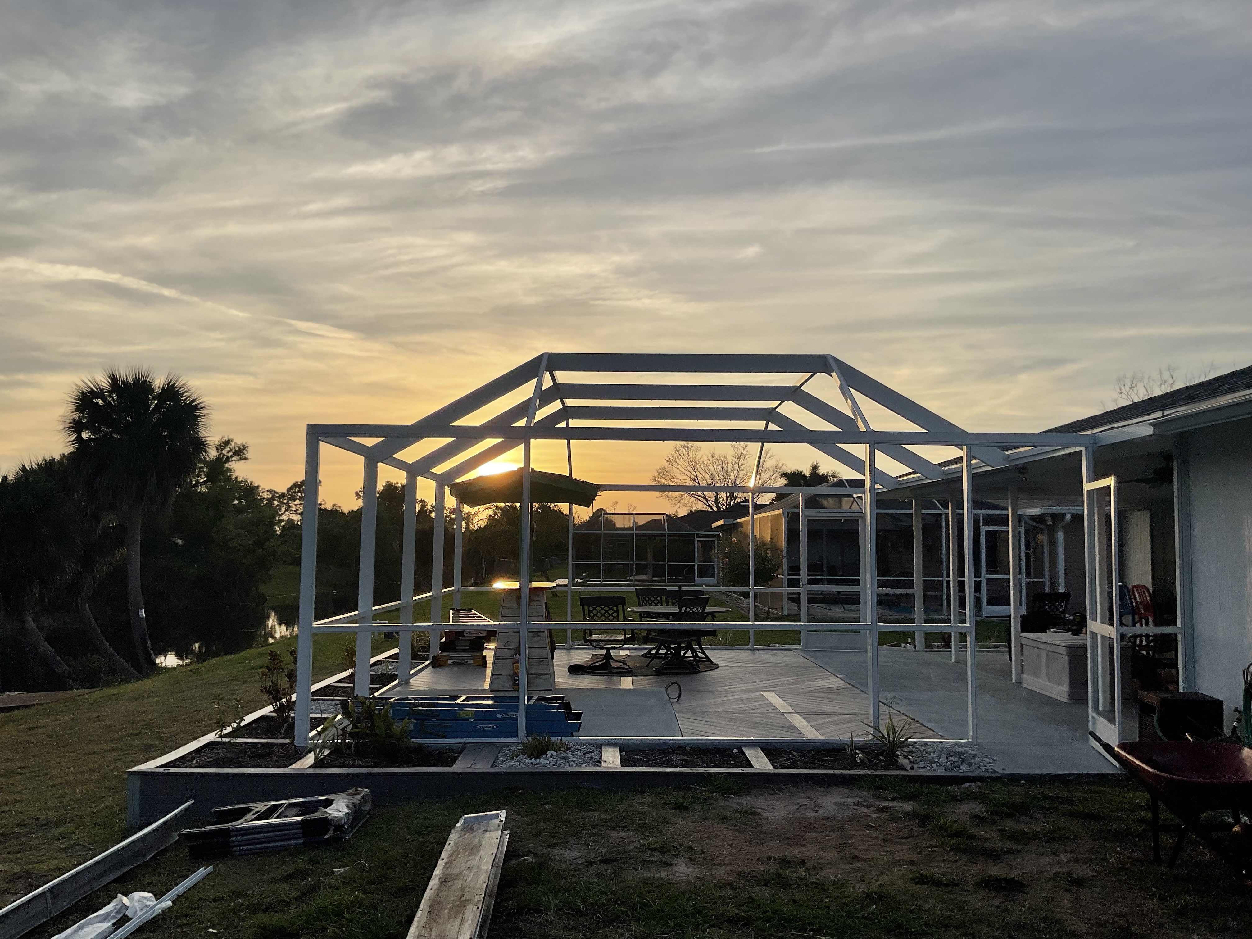White aluminum pool screen enclosure frame under construction at sunset, gable roof structure, concrete paver deck, South Florida lakefront backyard — AB Aluminum & Screens
