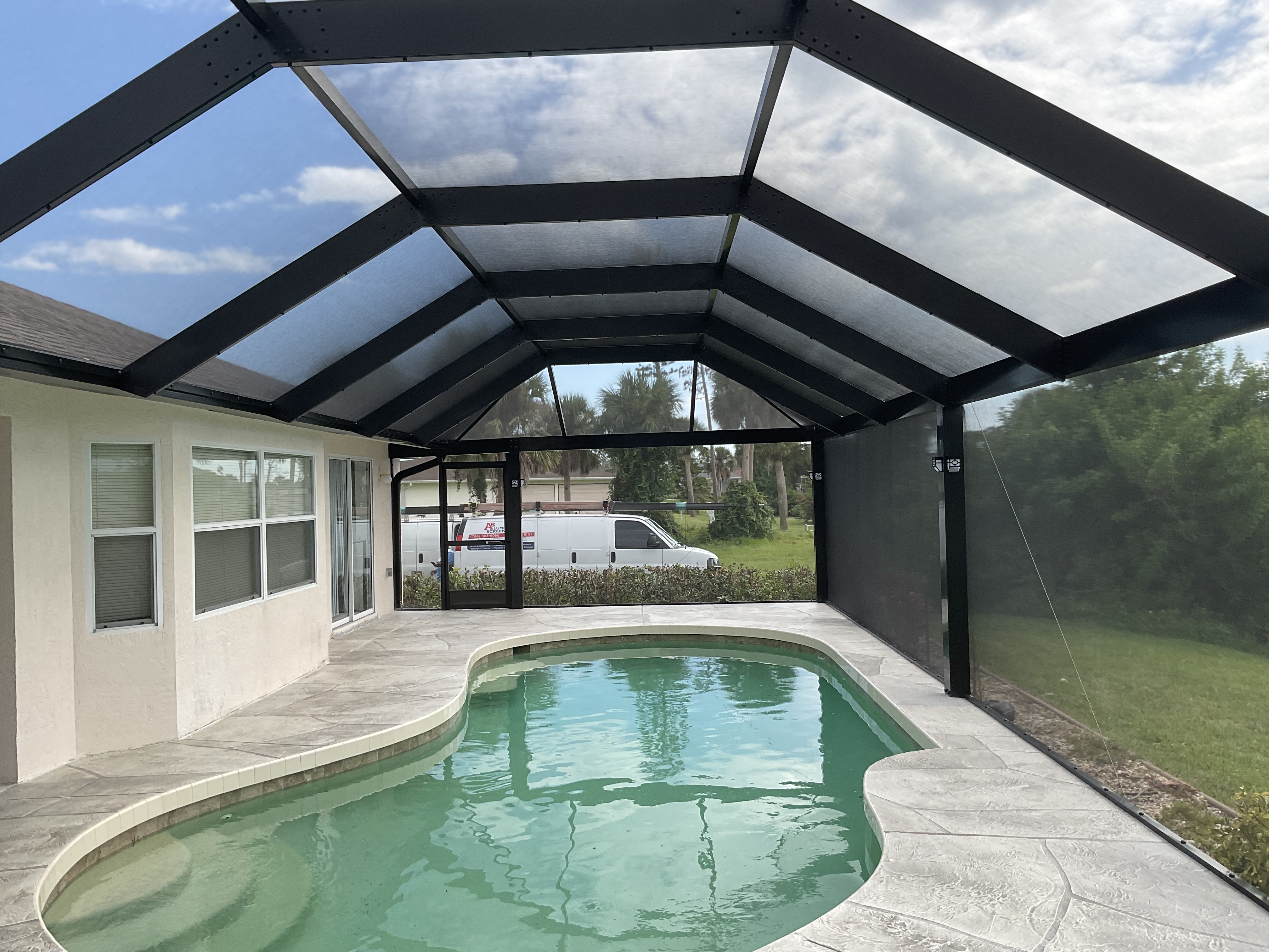 Dark bronze aluminum pool screen enclosure with gable roof, freeform pool, stamped concrete paver deck, single-story white stucco home, South Florida — AB Aluminum & Screens
