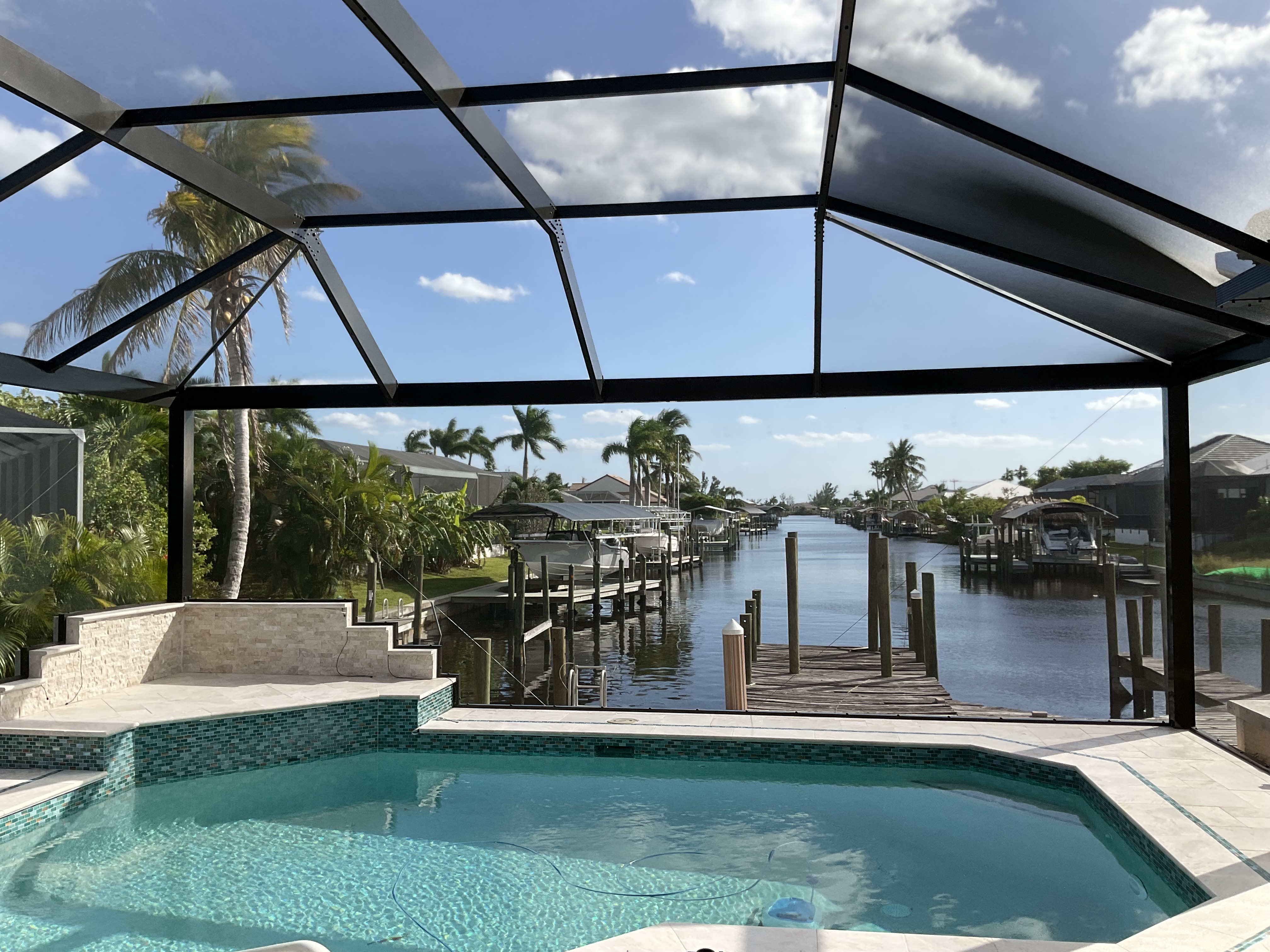 Dark bronze aluminum pool screen enclosure interior, freeform pool with elevated spa, waterfront canal view with boat docks and palm trees, South Florida — AB Aluminum & Screens