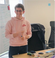 Personne souriante en sweat rose debout dans un bureau devant un tableau blanc et un bureau.