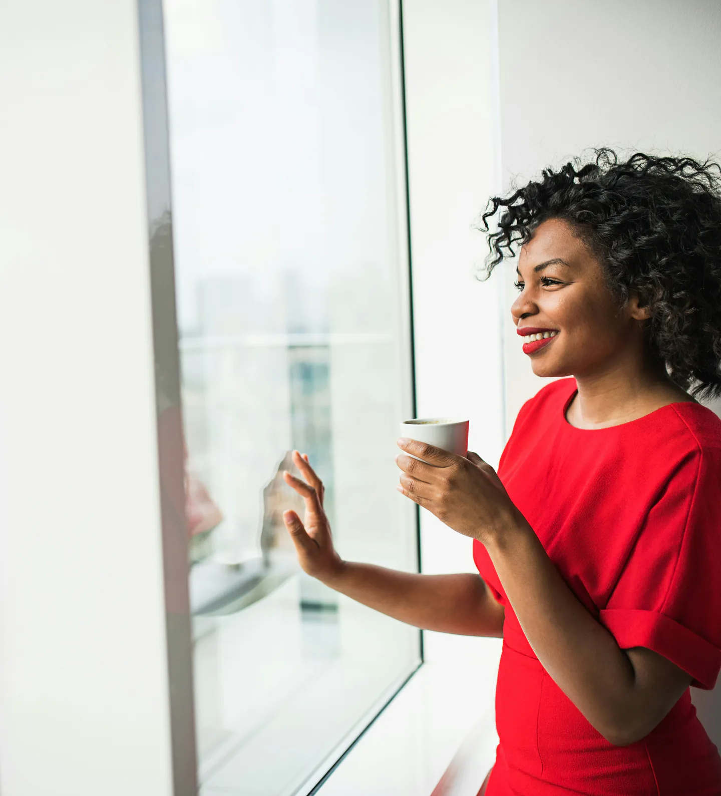 Une femme souriante, une tasse à la main, regarde par la fenêtre de son bureau.