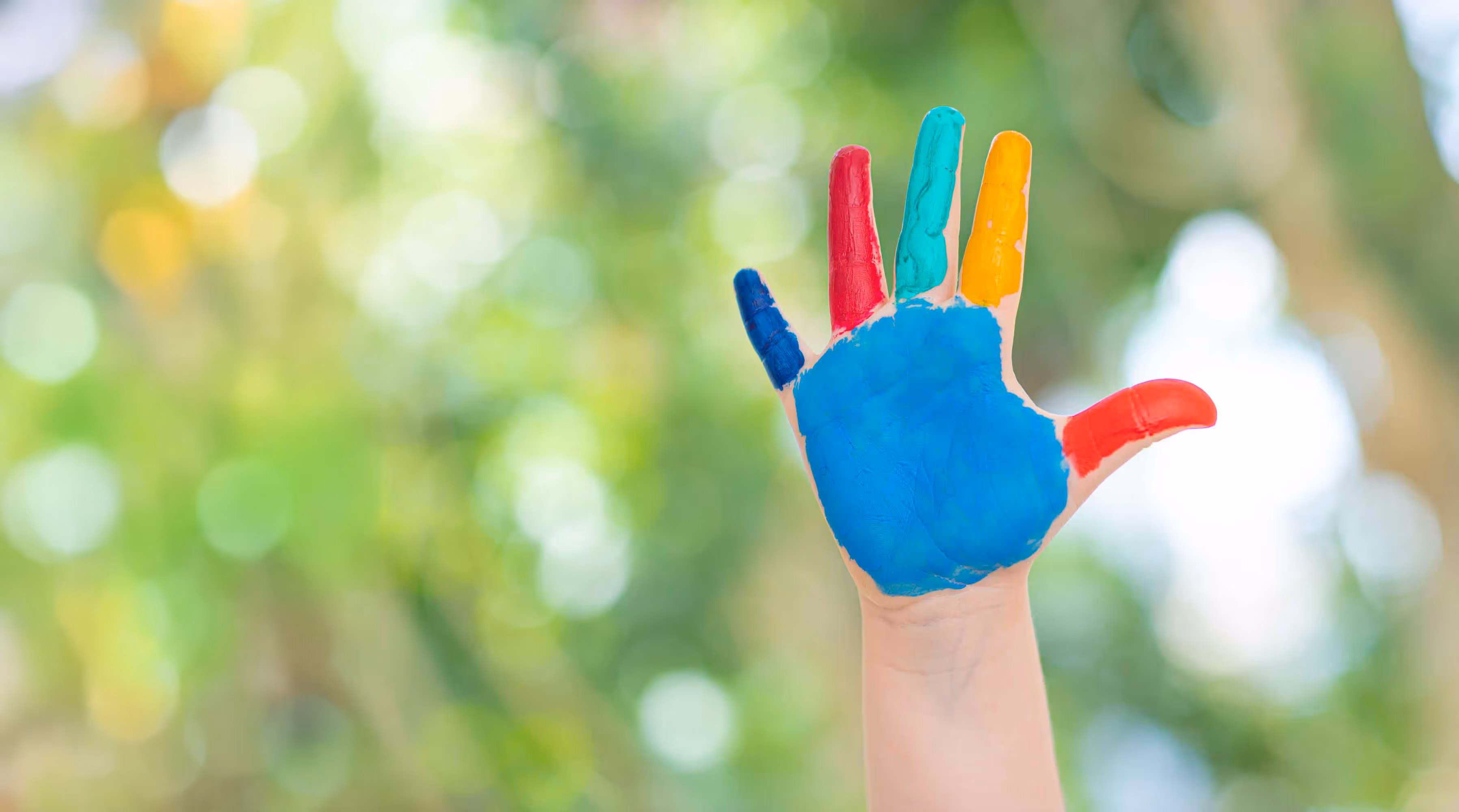 A hand with colorful paint on fingers and palm is raised against a blurred green background.