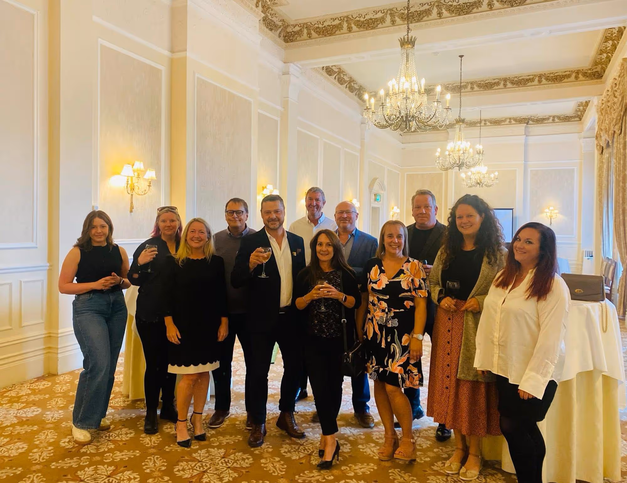 A group of people stand together in a formal room, holding glasses and smiling at the camera.