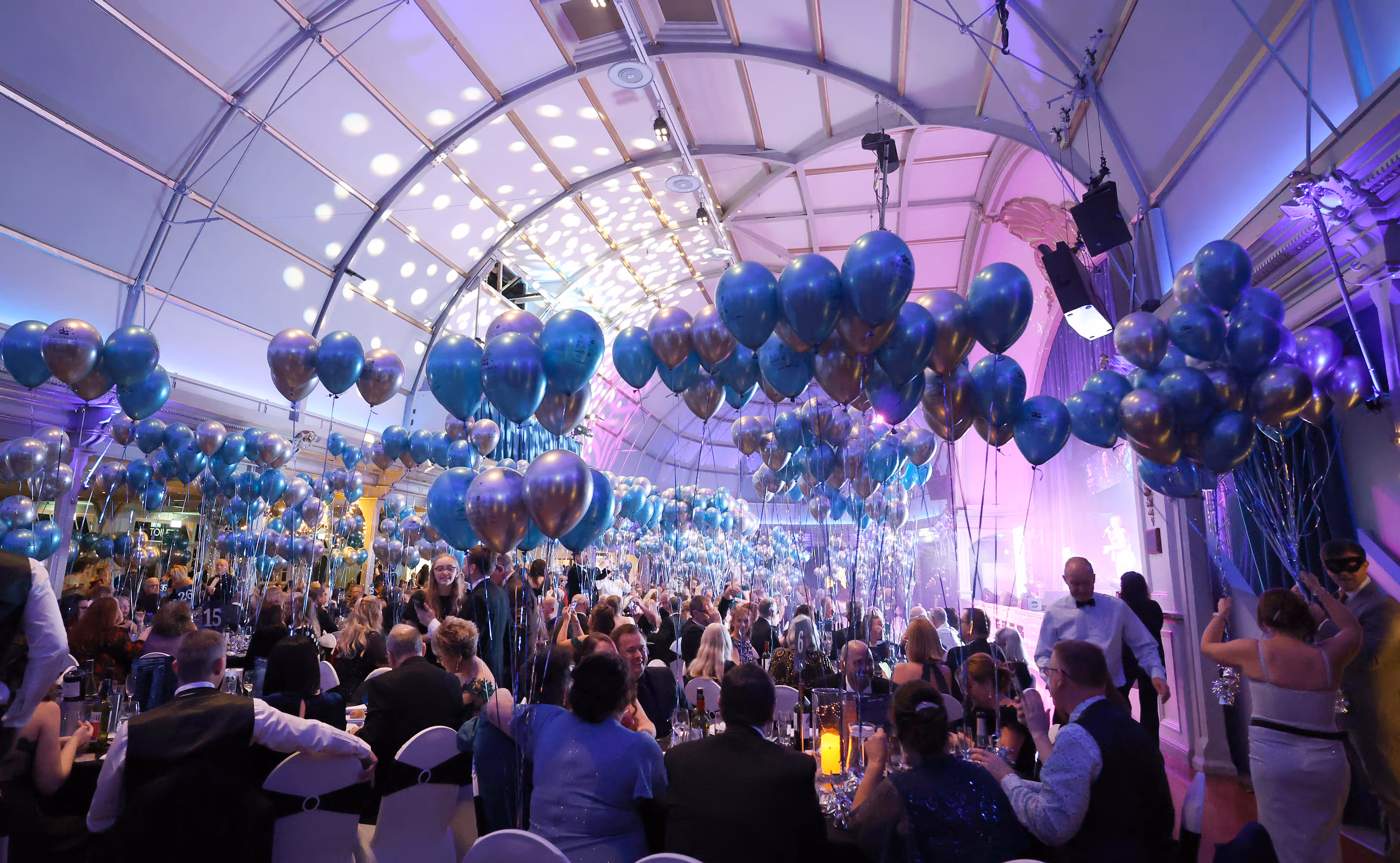 A large event hall filled with people seated at tables, with blue balloons hanging overhead.
