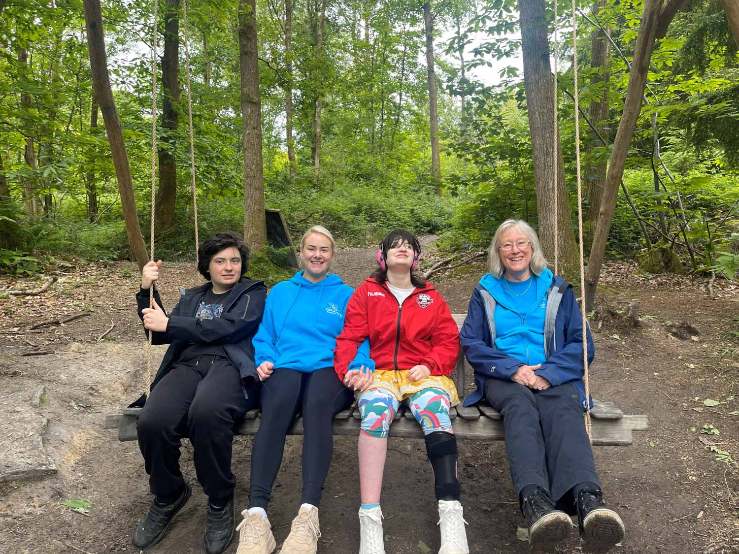 Four people sit together on a wooden swing in a forest, smiling and holding hands.