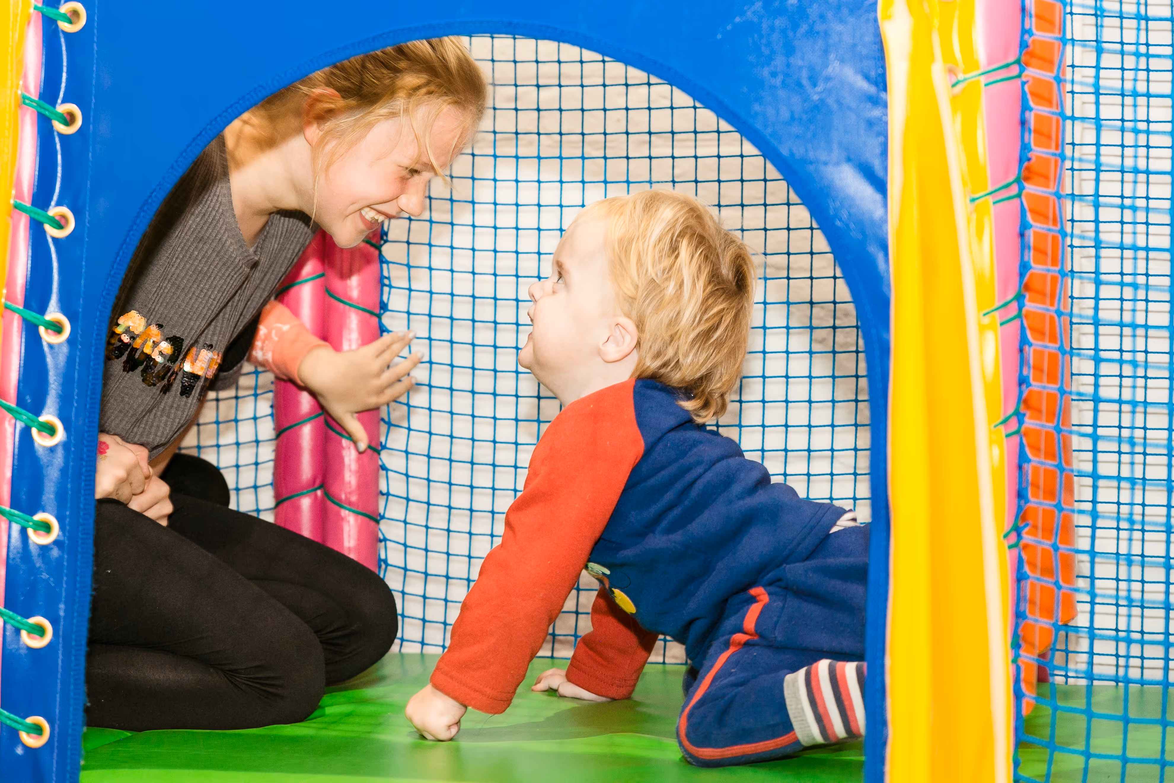 Child in a blue onesie and adult smiling at each other inside a colorful play area.