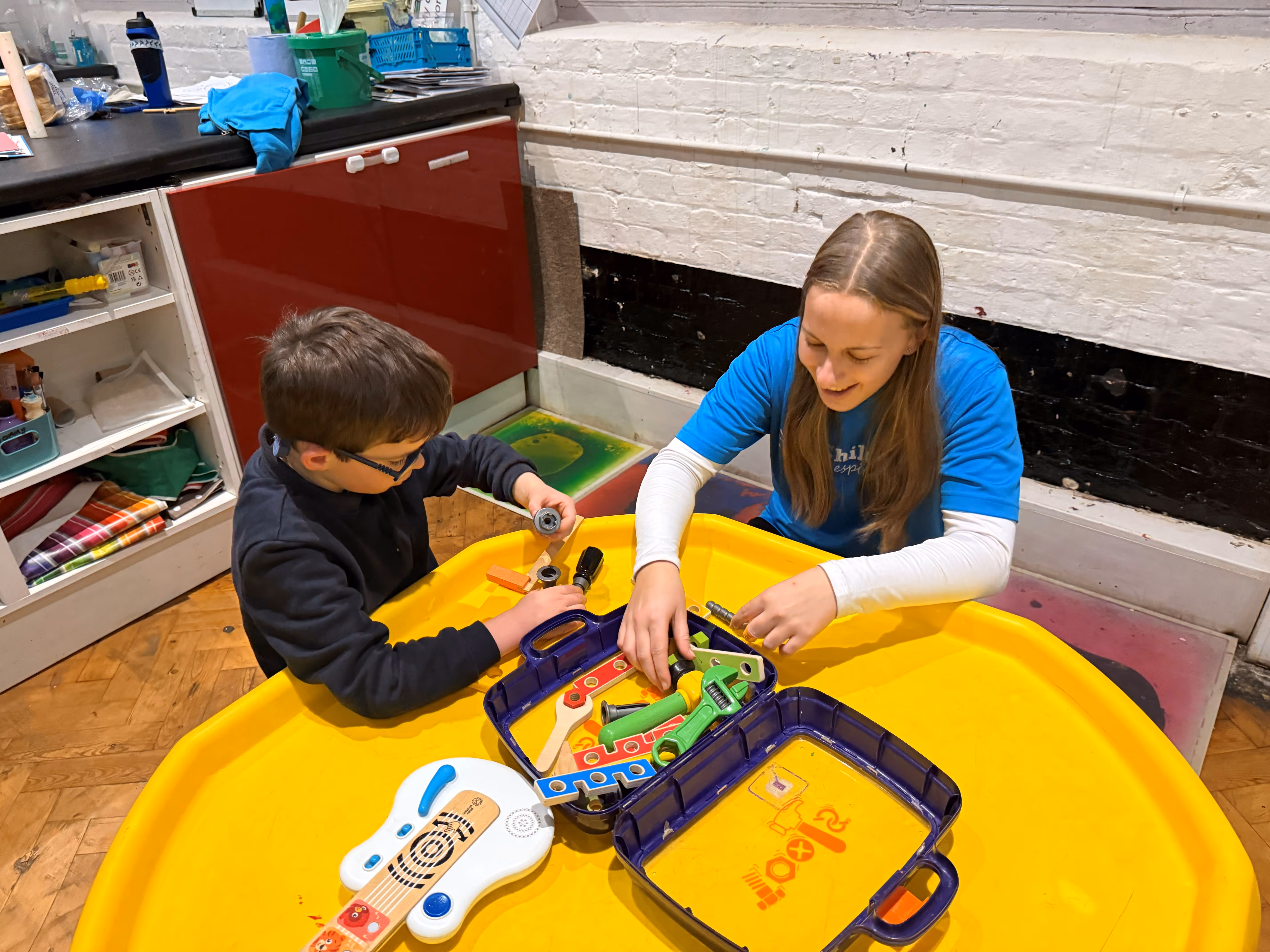 A woman and a child play with colorful toy tools on a yellow table.