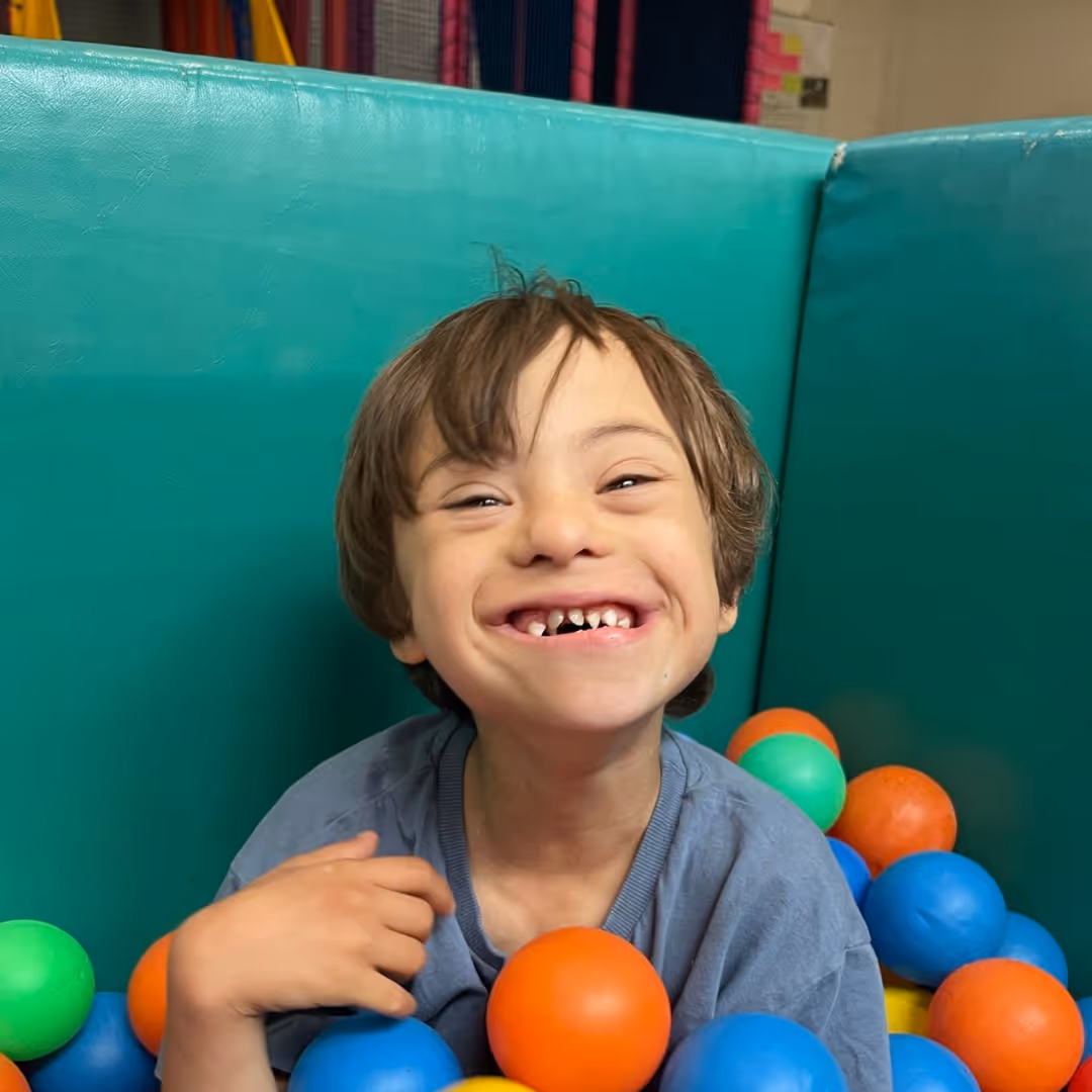 A smiling child with missing front teeth sits in a colorful ball pit.