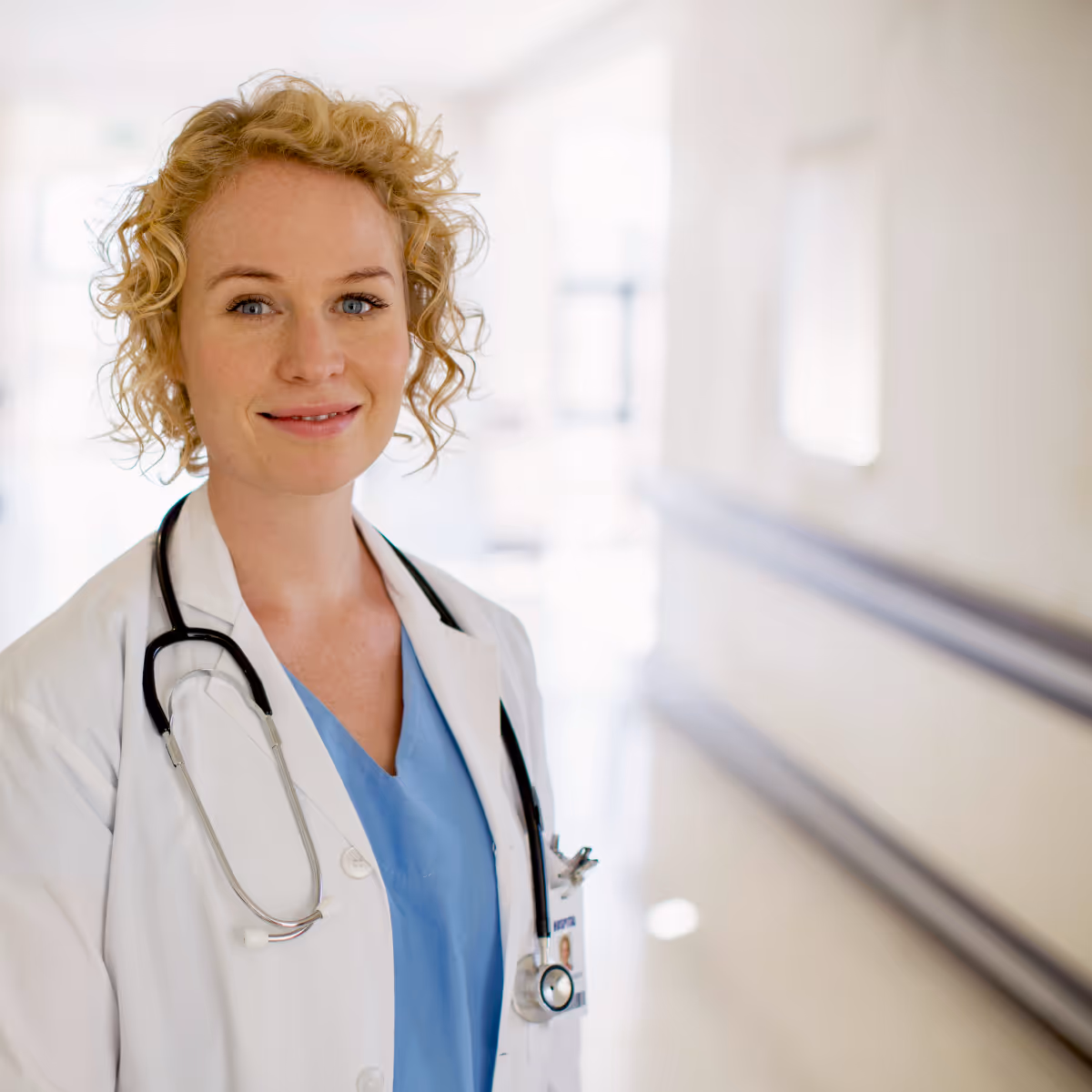 Kvinnlig blond läkare med stetoskop och blå sjukhuskläder ler i sjukhuskorridor.
