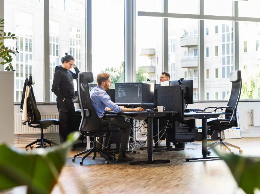Tre personer samarbetar i ett modernt kontorslandskap med stora fönster och datorer på skrivbordet.