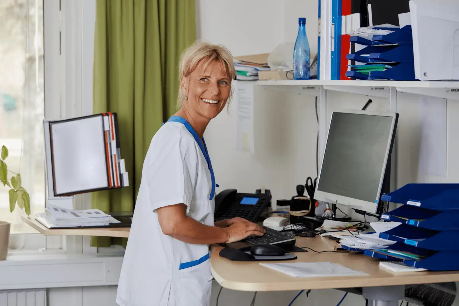 Leende sjuksköterska vid ett stående skrivbord med dator och organiserade kontorsmaterial.