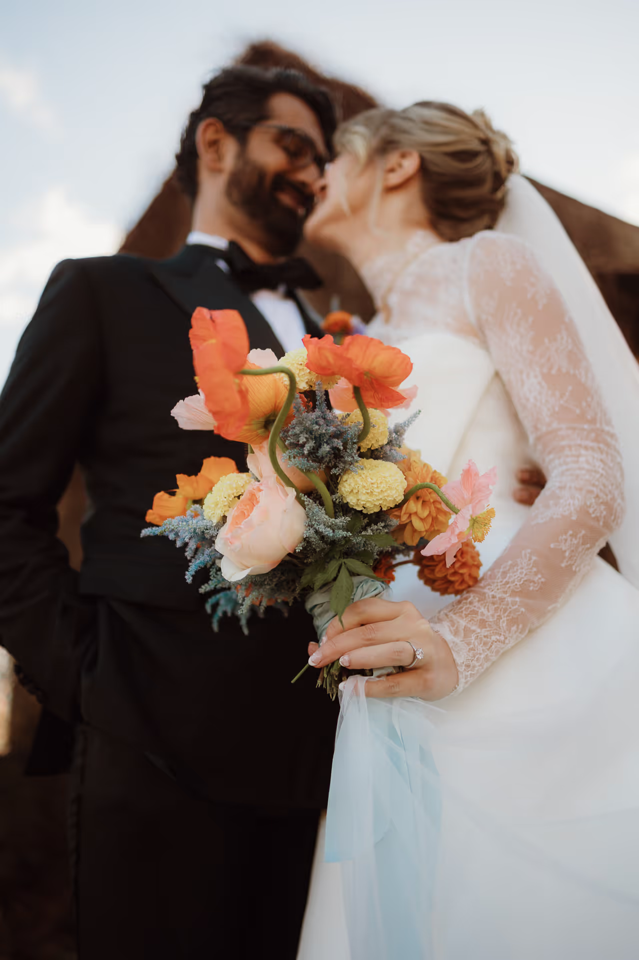 Groom kissing the bride's forehead as she holds an orange and coral bouquet