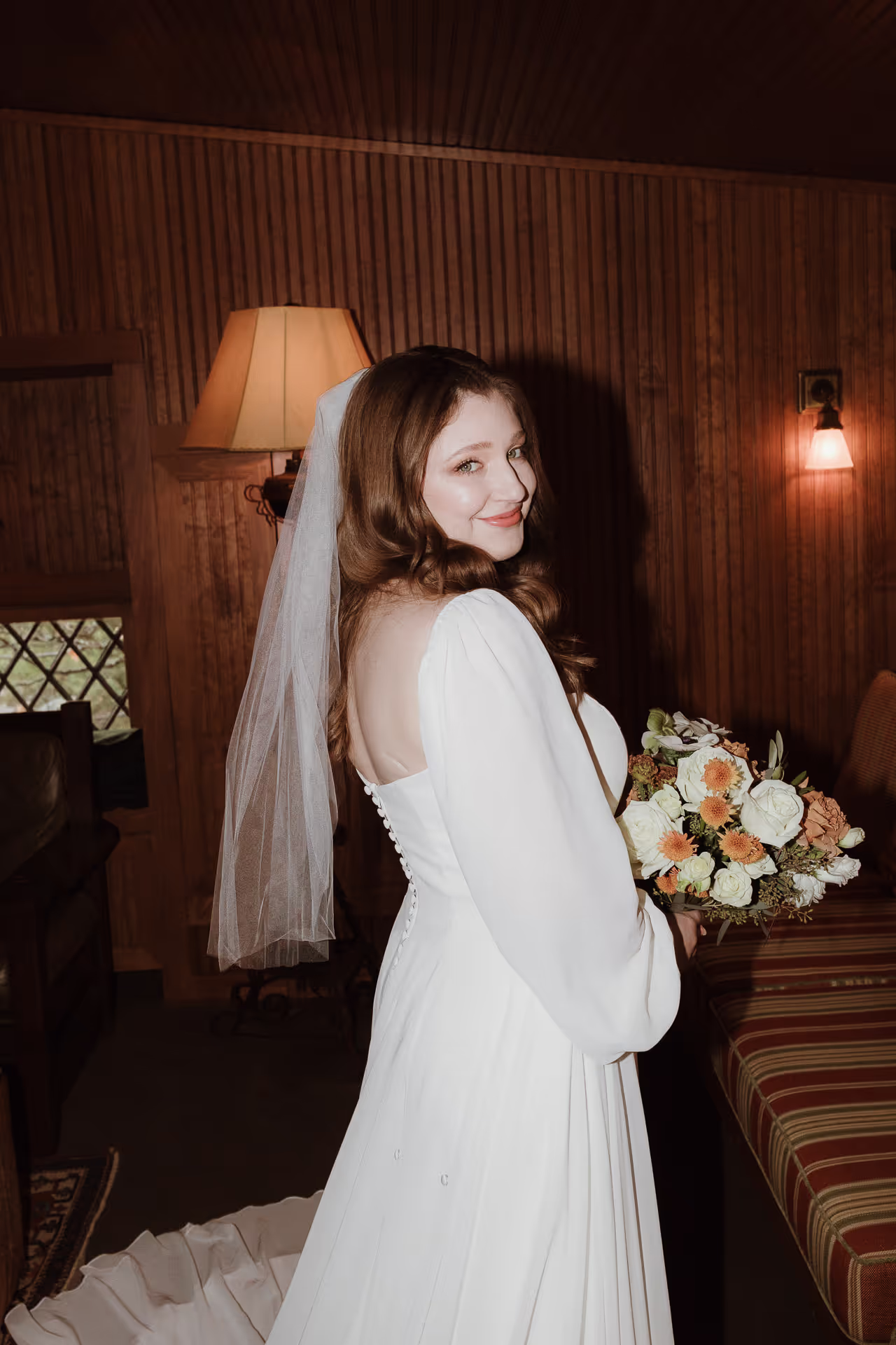Bride with a veil and bouquet smiling over her shoulder in a wood-panelled room at Deer Mountain Inn
