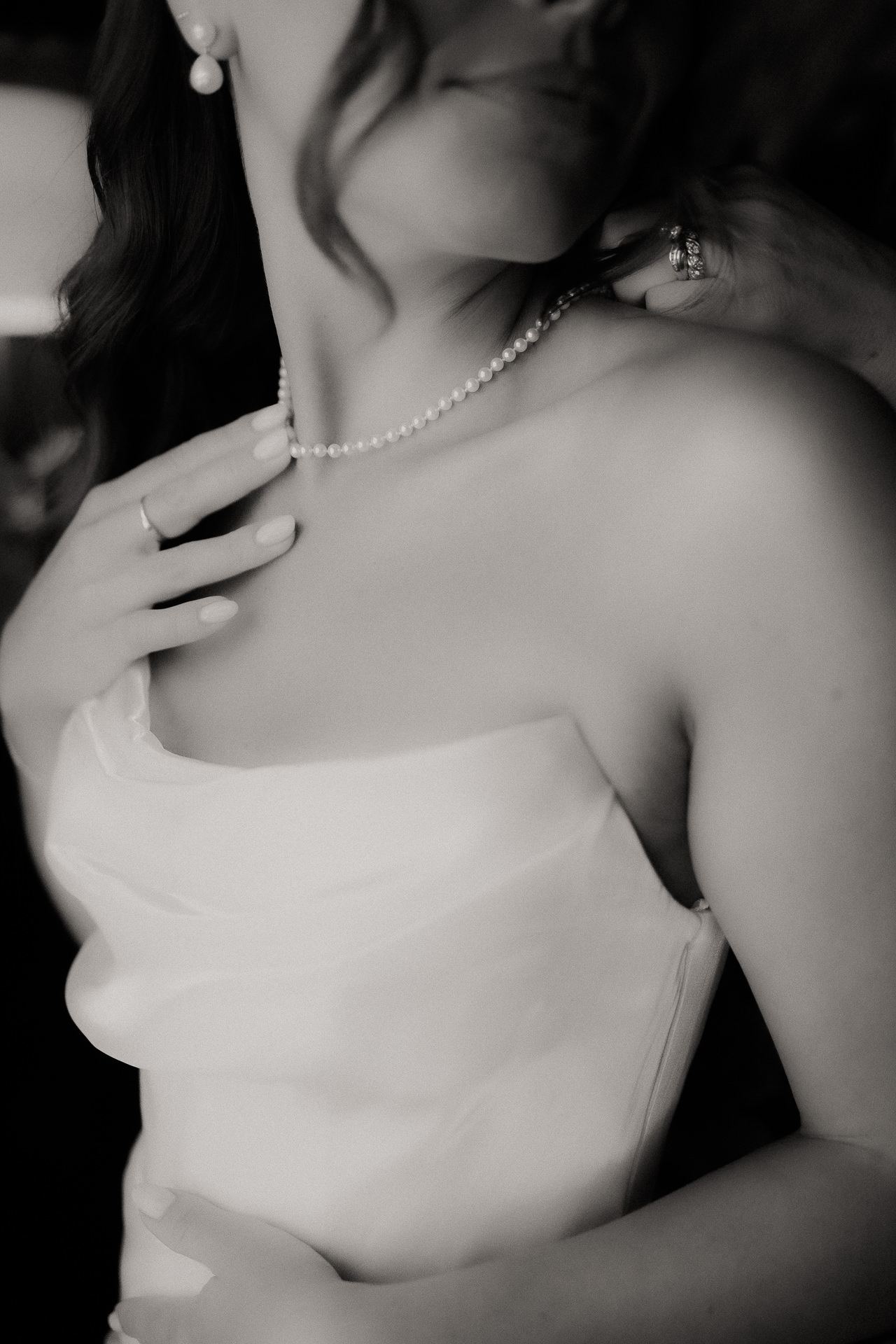 Black and white photograph of a bride fastening a pearl necklace while getting ready