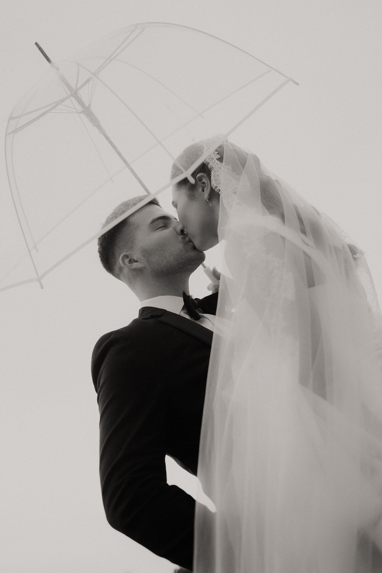 Black and white photograph of a bride and groom kissing under a clear umbrella in the rain