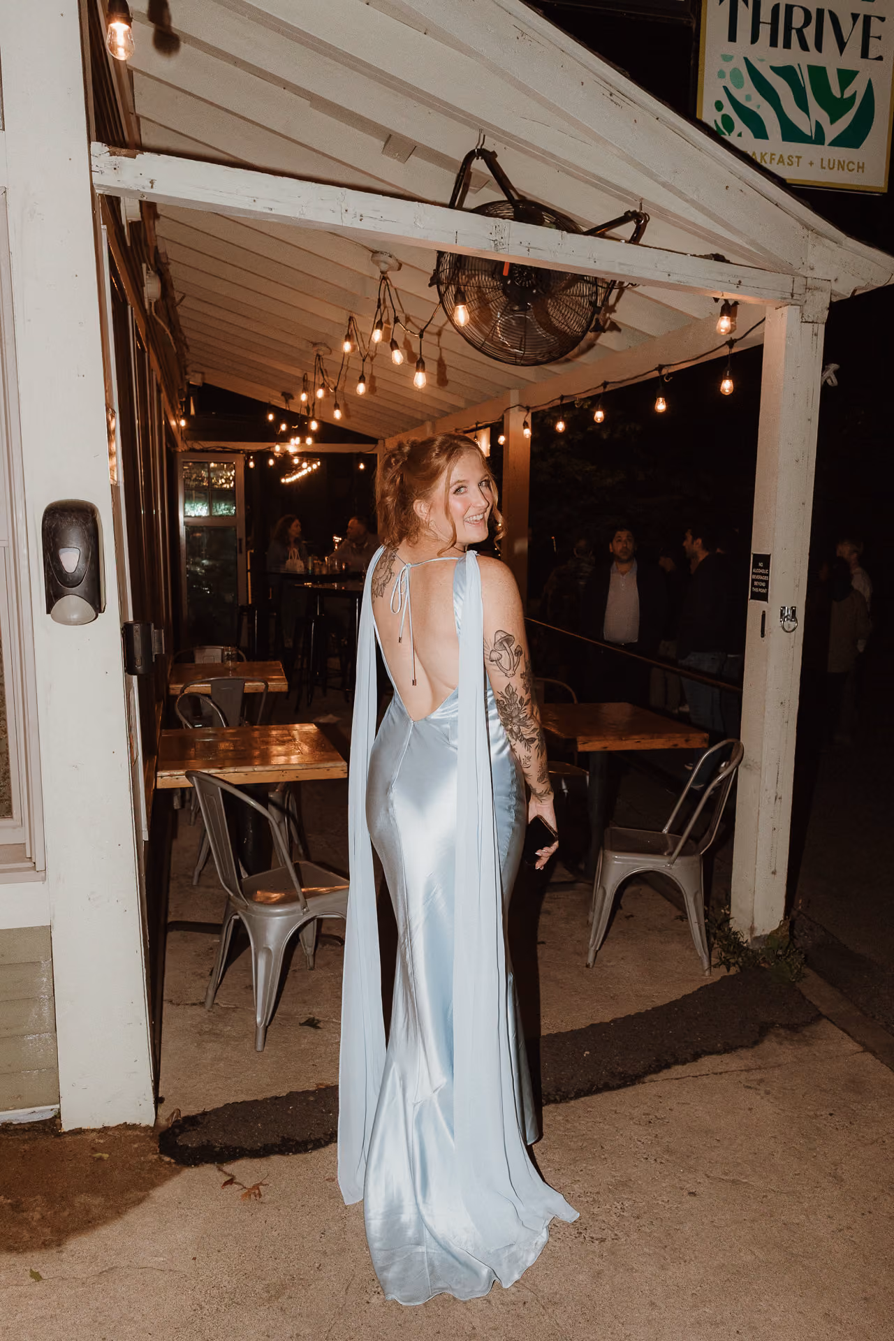 Bride in a pale blue satin gown standing in a string-lit restaurant doorway at a welcome party