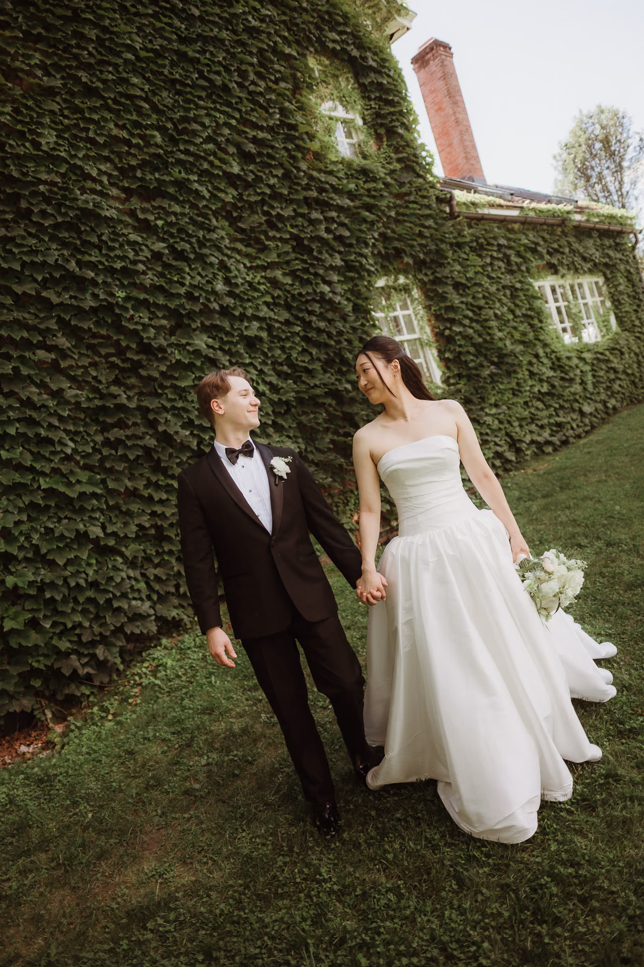 Bride and groom walking hand in hand beside an ivy-covered house at Windrift Hall